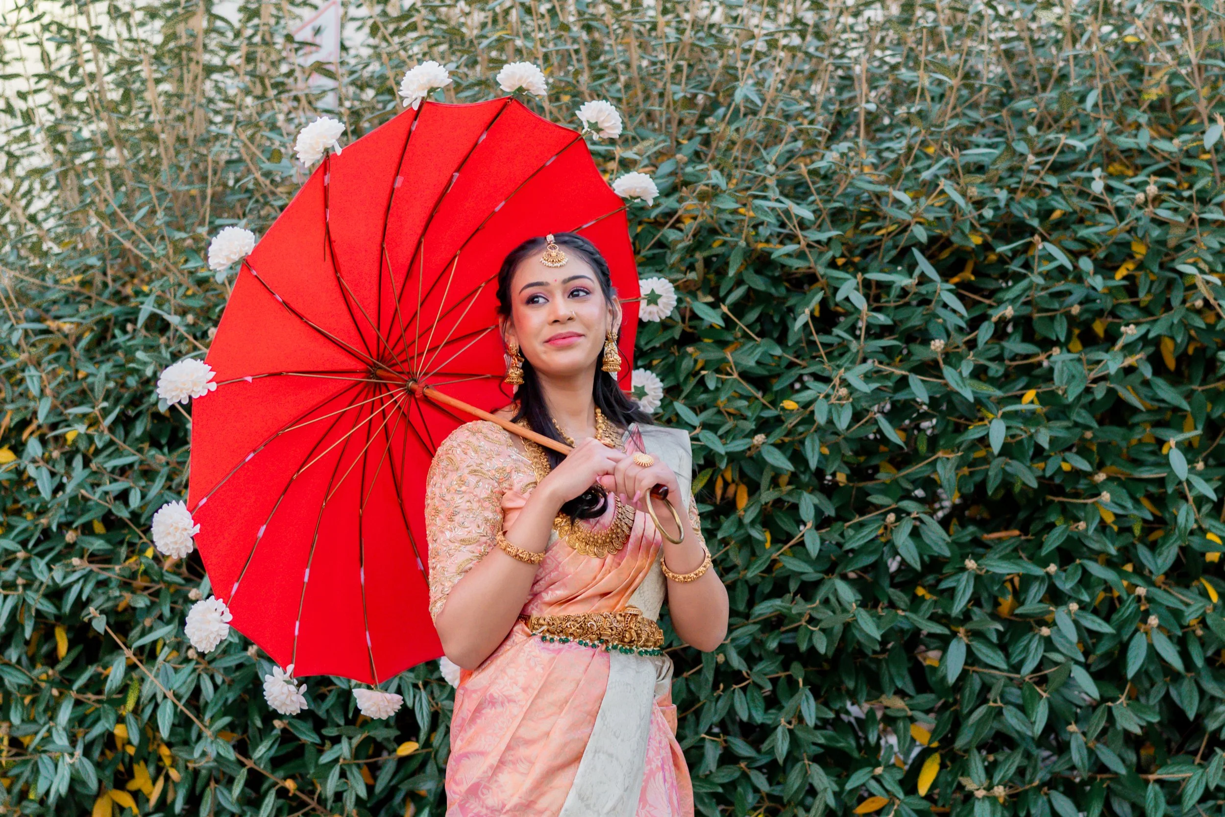 A woman dressed in traditional Indian attire holding a red parasol decorated with white flowers, standing in front of a lush green hedge.