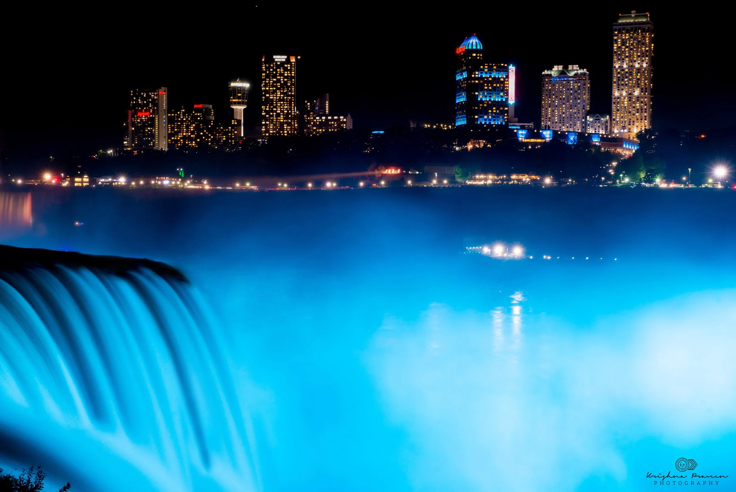 Night view of the city skyline with illuminated buildings, viewed from behind a waterfall with water cascading into a pool below.