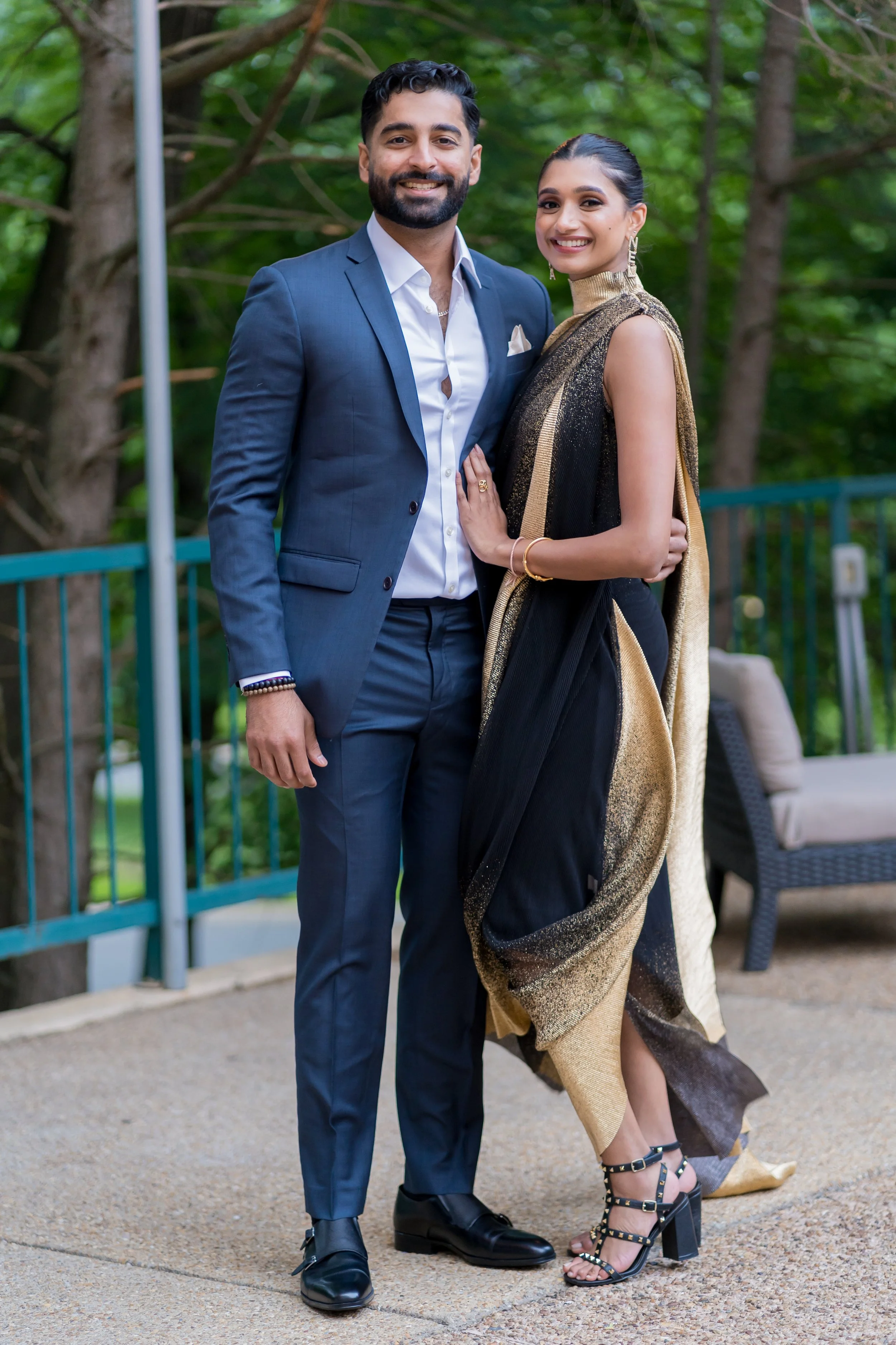 A smiling man and woman dressed in formal attire standing outdoors with a green wooded background. The man wears a navy blue suit with a white shirt and black shoes, and the woman wears a black and gold dress with high heels, jewelry, and has her hai