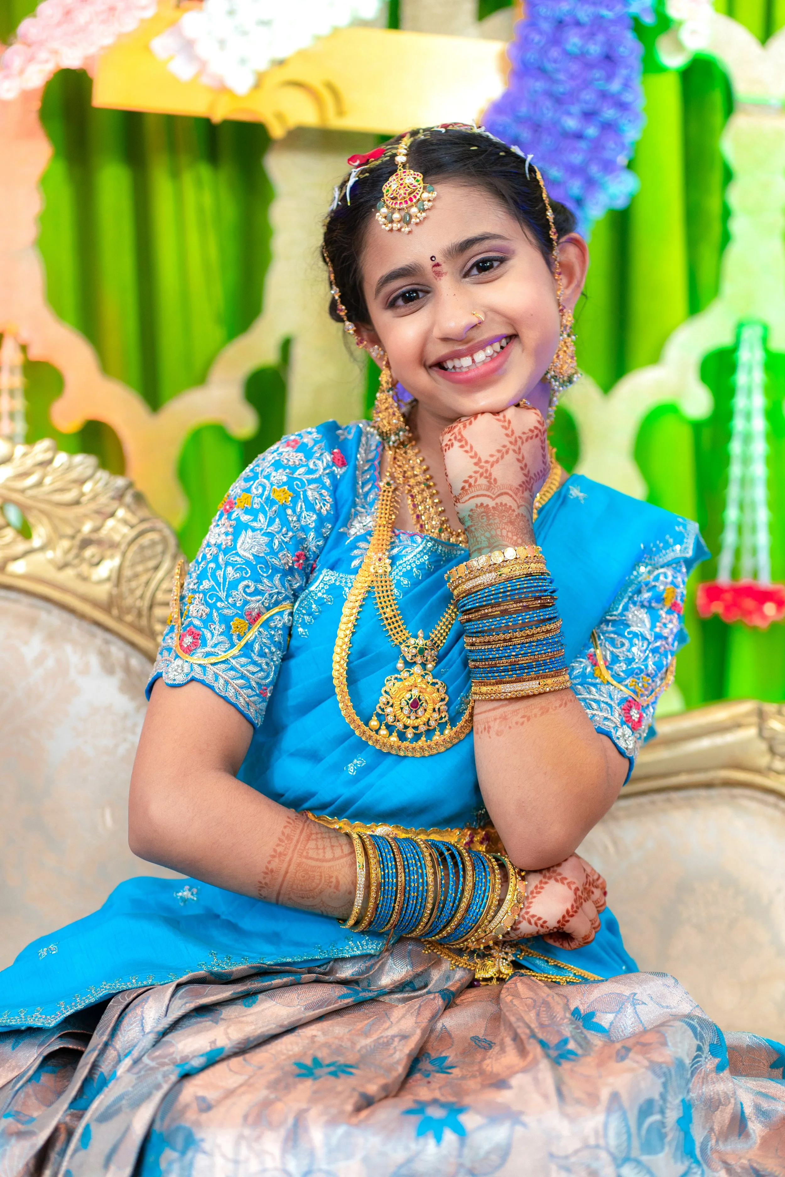 A young girl in traditional Indian attire, smiling with her head resting on her hand, adorned with jewelry and henna tattoos, in front of a colorful backdrop with green and purple decorations.