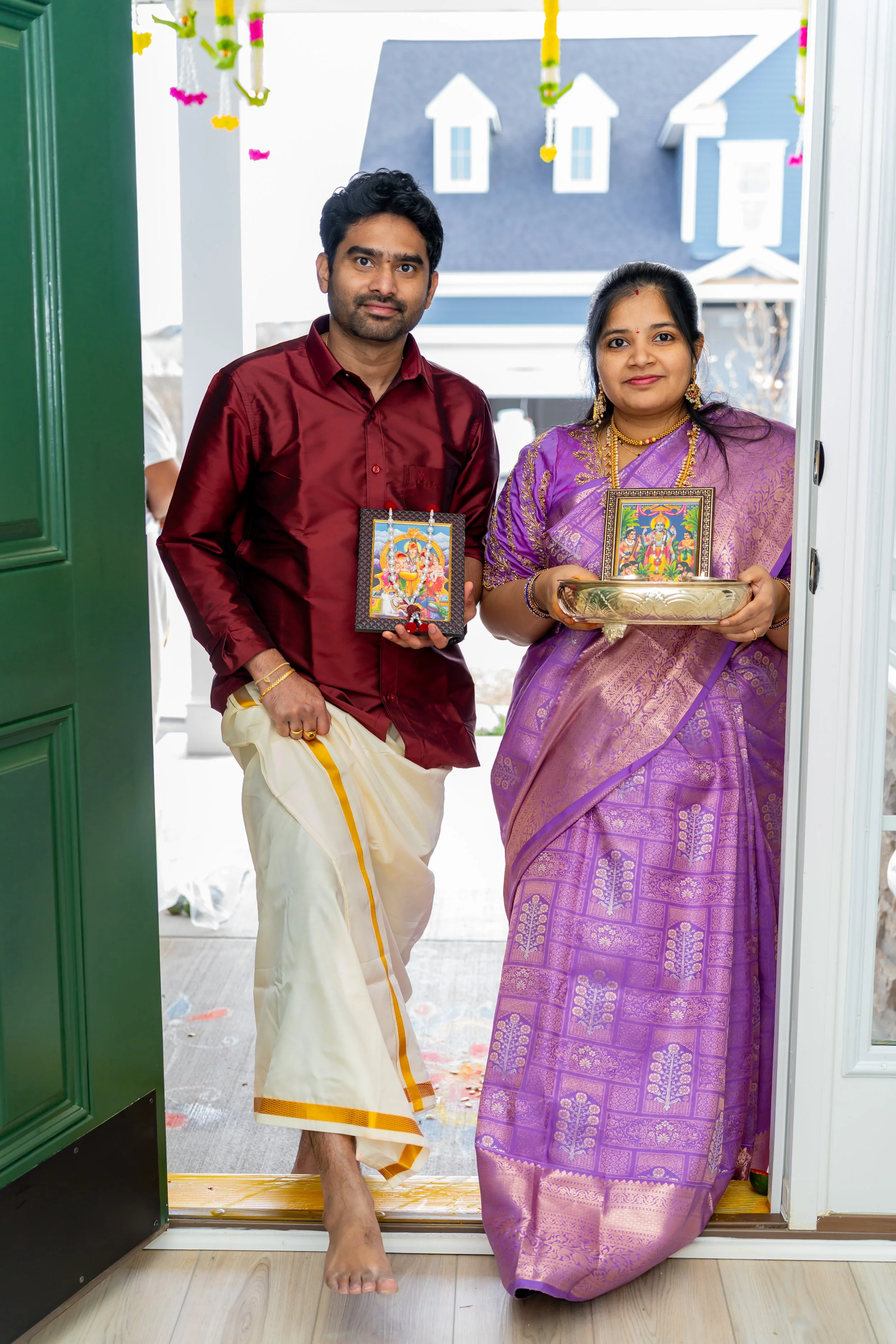 Indian man and woman dressed in traditional attire standing at the door of a house, holding images of Hindu deities during a religious celebration or festival.