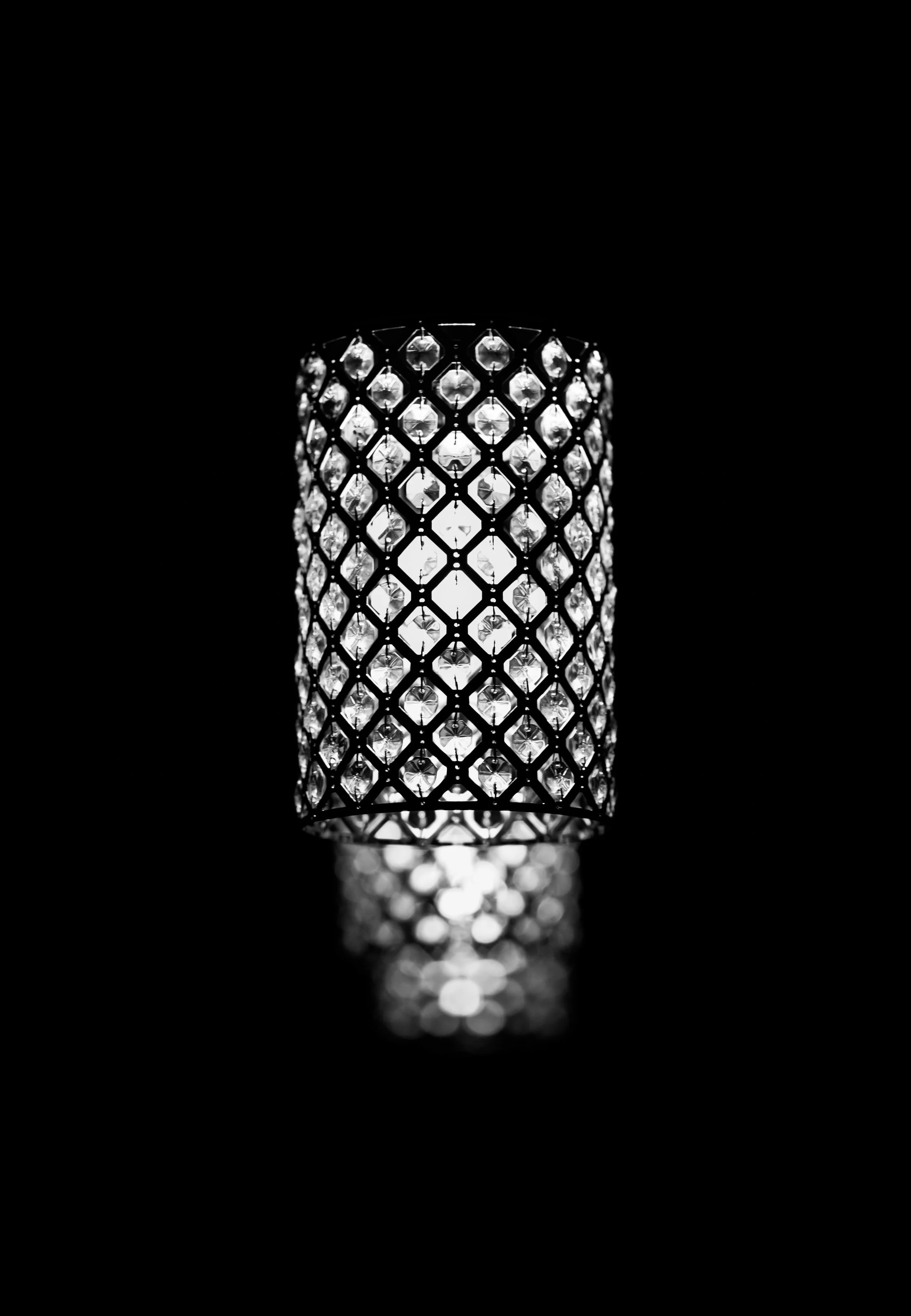 A black-and-white photo of a cylindrical lampshade with a lattice pattern of cutouts and decorative crystals inside, emitting light and casting a pattern on the surface below.