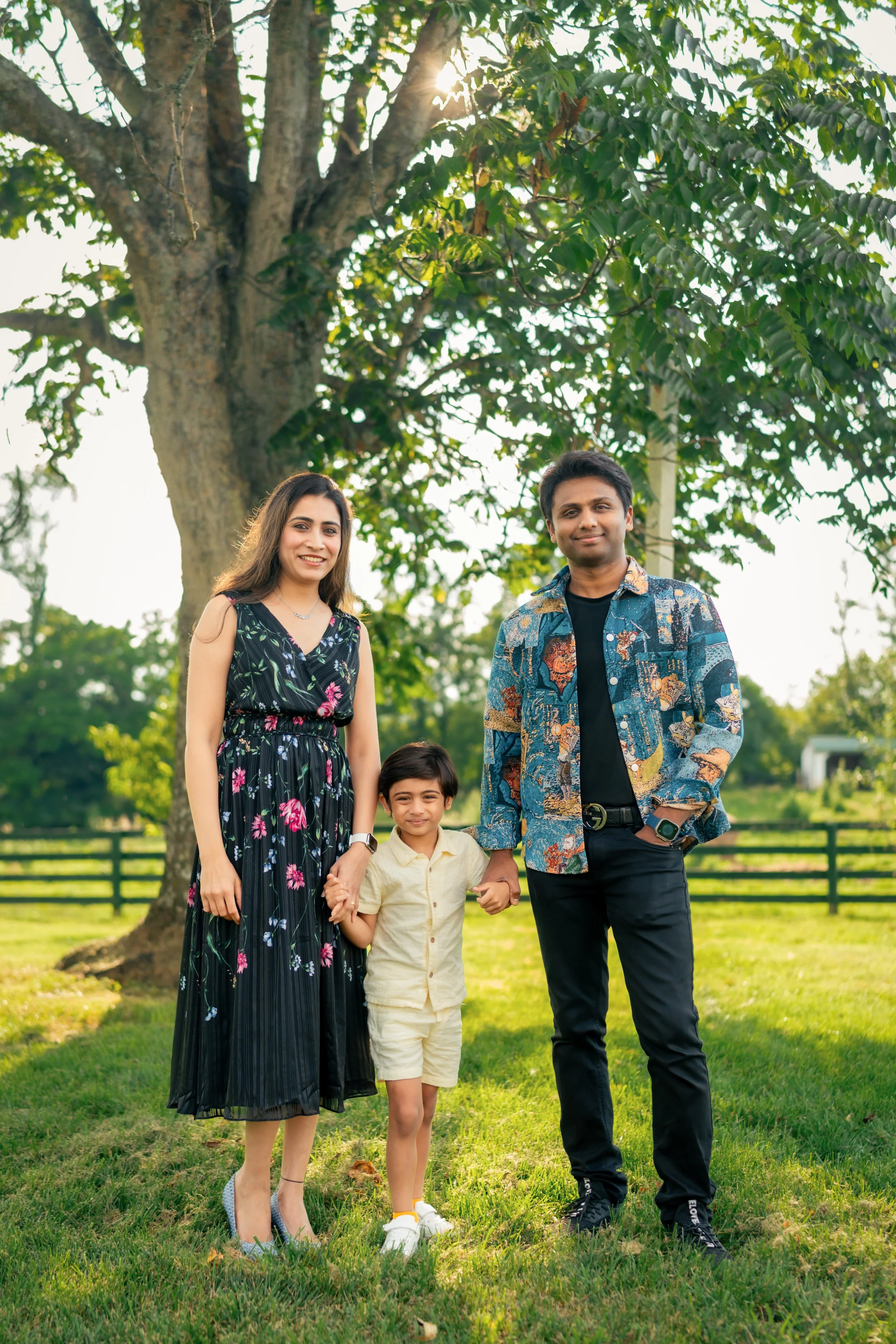 A family of four standing outdoors in front of a large tree with sunlight shining through the branches, all smiling. The family includes a woman in a floral dress, a man in a colorful patterned shirt, a young boy in a light yellow shirt, and a young 