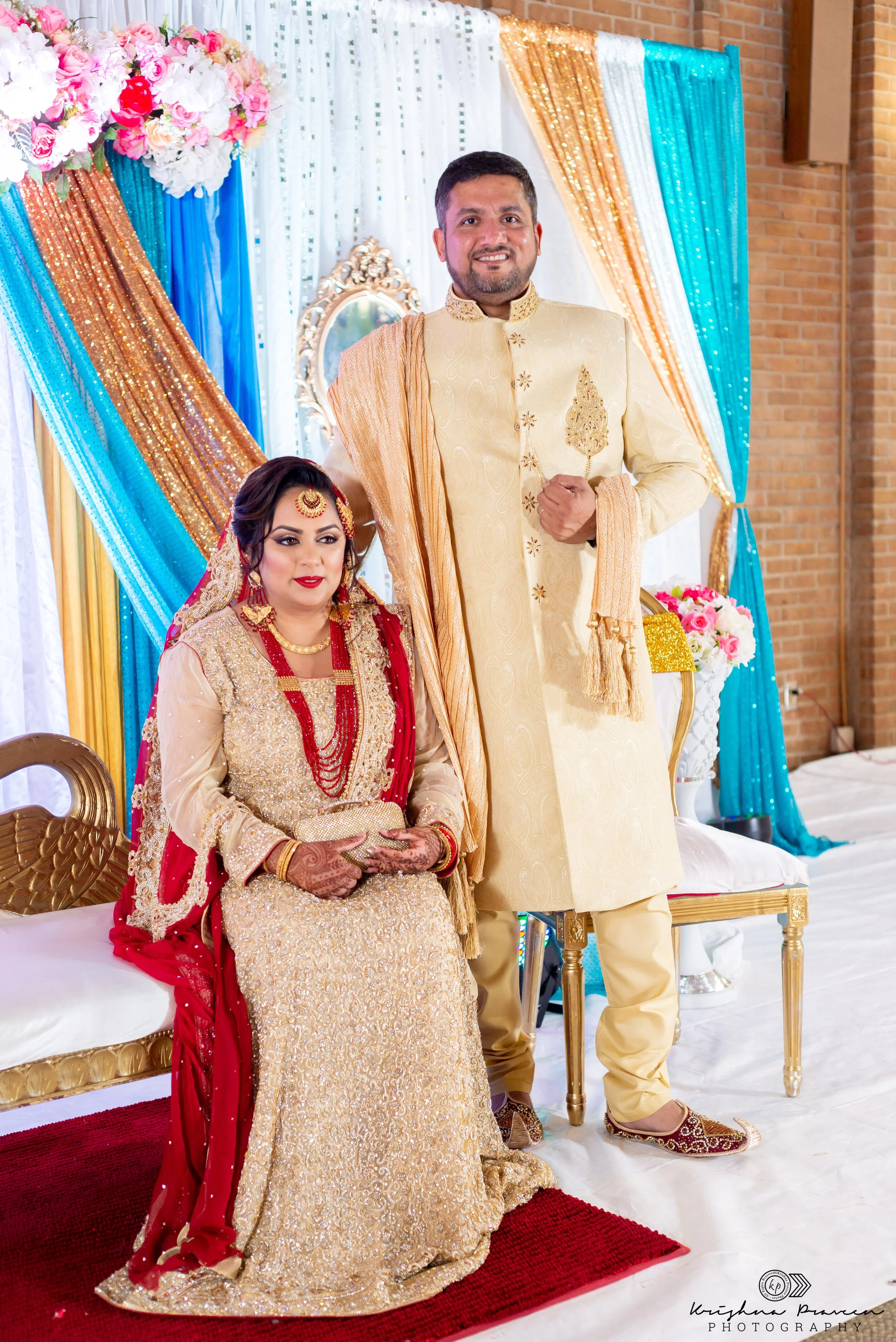 A bride and groom at their wedding, dressed in traditional South Asian attire. The bride is seated on a bench, wearing a gold embroidered dress with a red dupatta and intricate jewelry. The groom stands next to her in a cream-colored sherwani with go