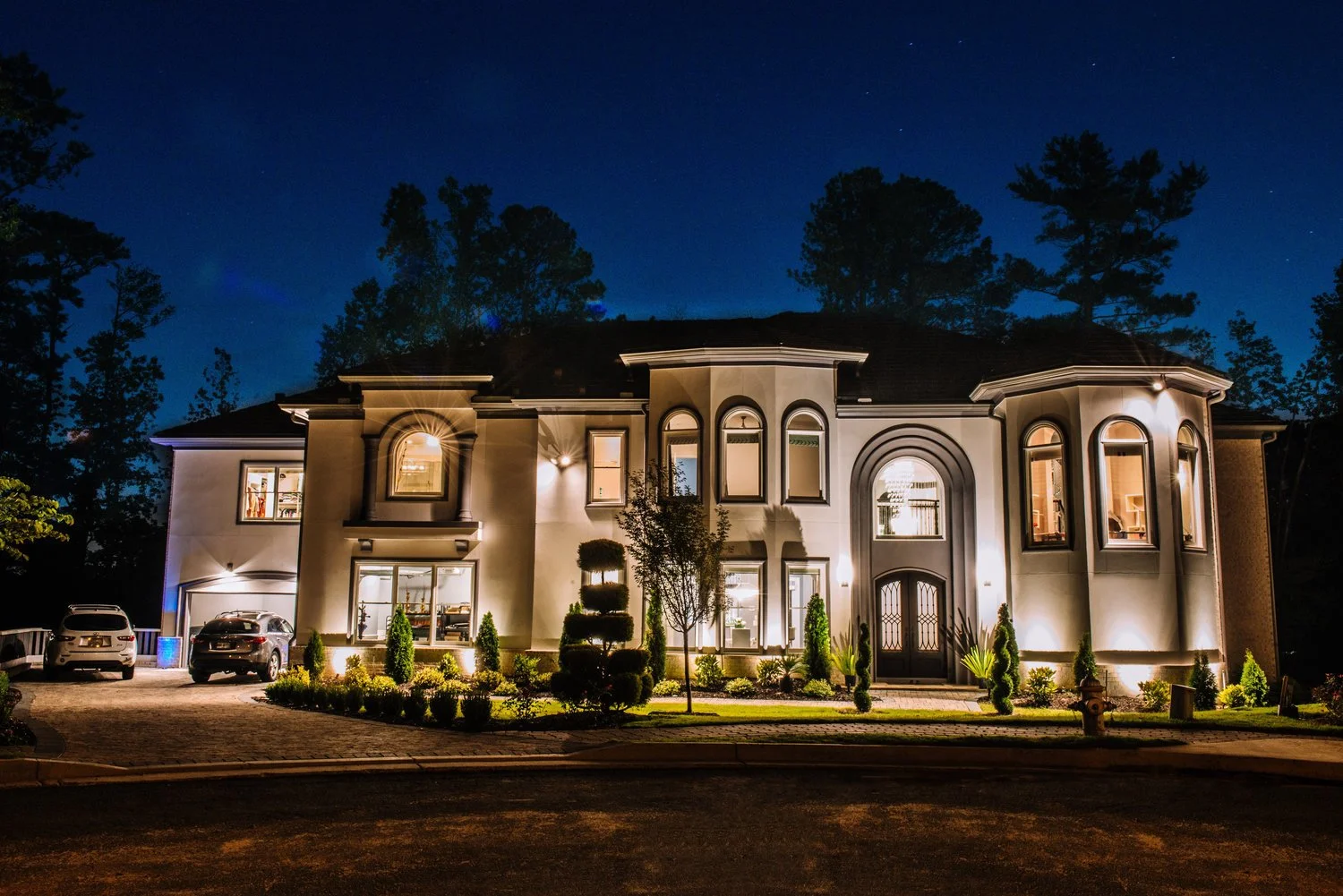 Large upscale house illuminated at night with trees in the background and cars parked in the driveway.