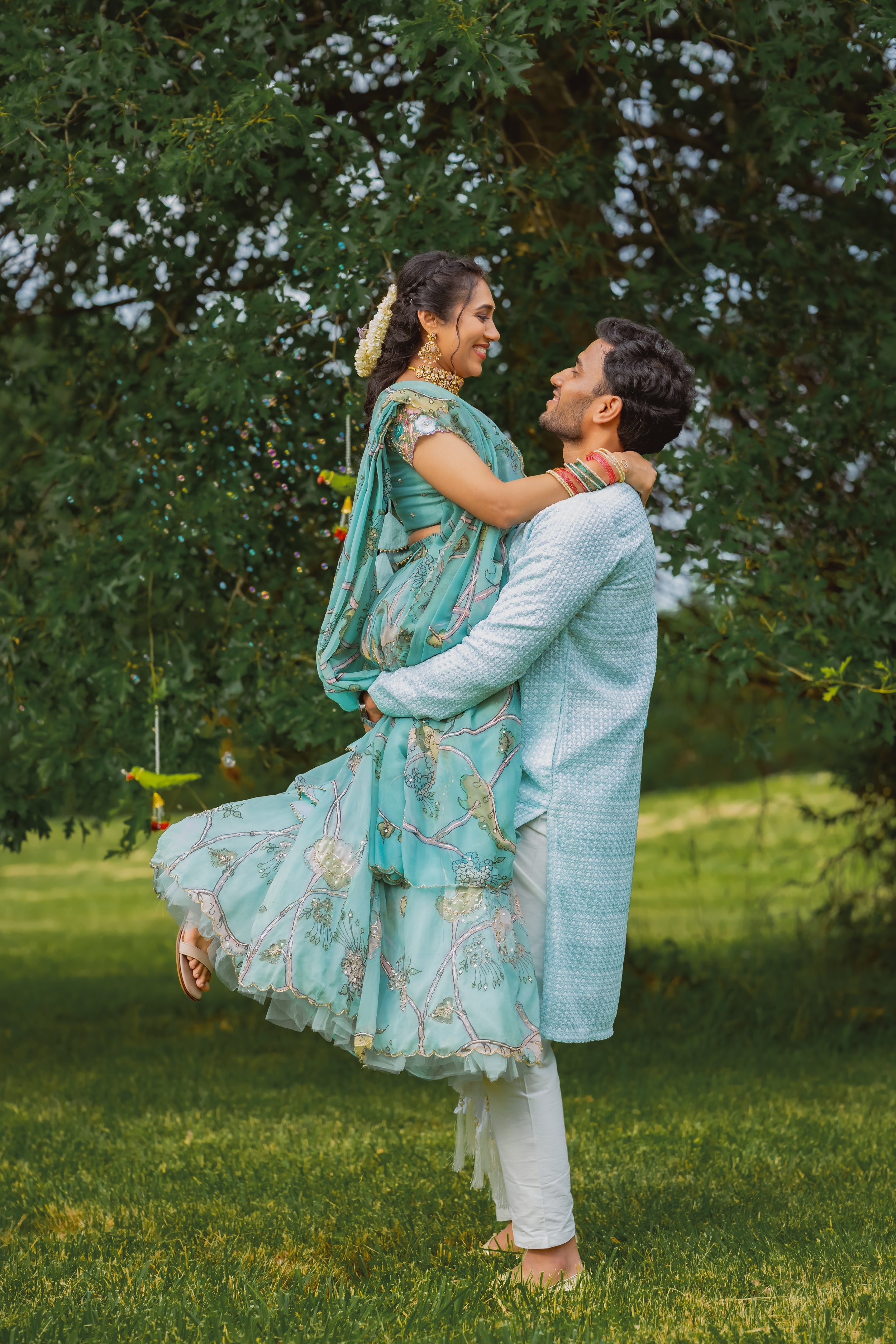 A couple dressed in traditional Indian attire, with the man lifting the woman in front of a green leafy tree, joyful expressions, outdoor setting.