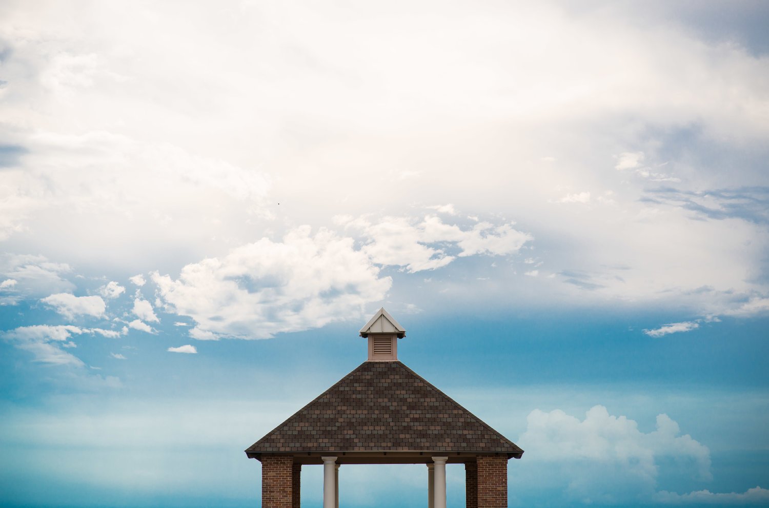 A small gazebo with a shingled roof and four white columns set against a cloudy sky.