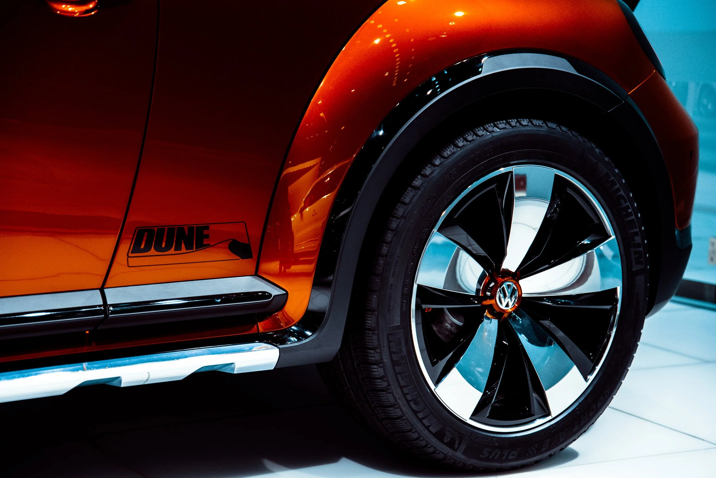Close-up of a shiny orange Volkswagen car's front fender and wheel, with a 'Dune' sticker near the bottom.
