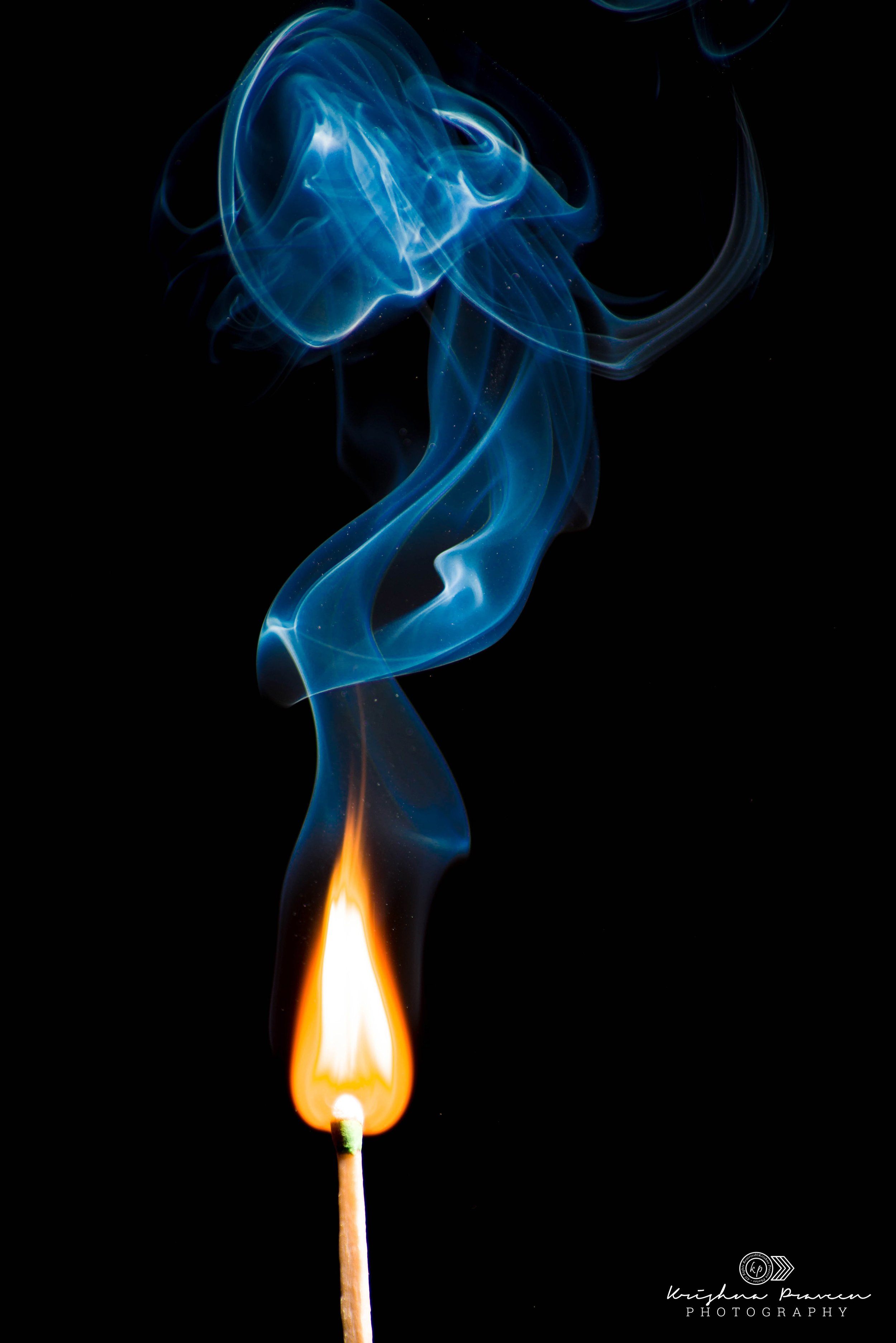 A matchstick ignited with a burning flame, producing blue, orange, and yellow smoke against a black background.