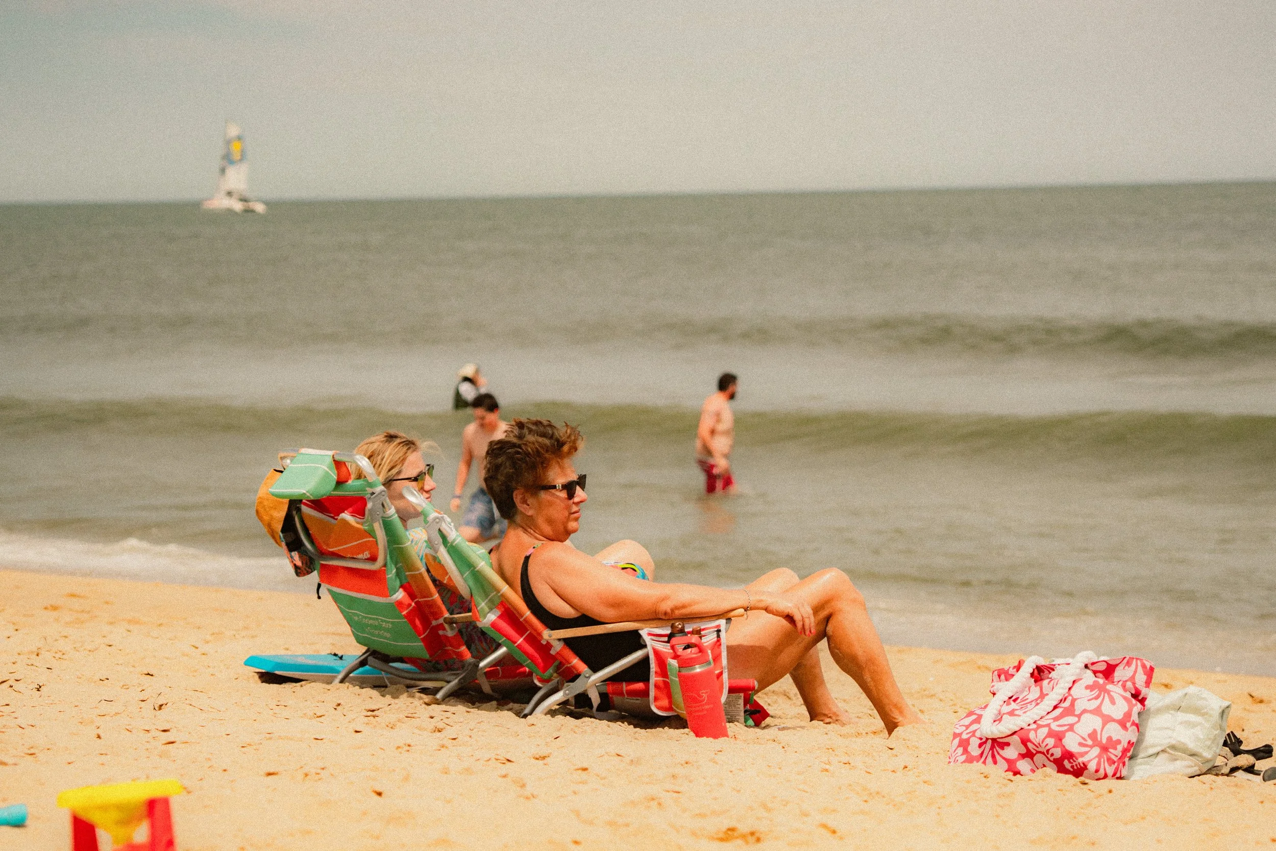 People sitting on beach chairs and relaxing on the sandy beach, with the ocean and a sailboat in the background.