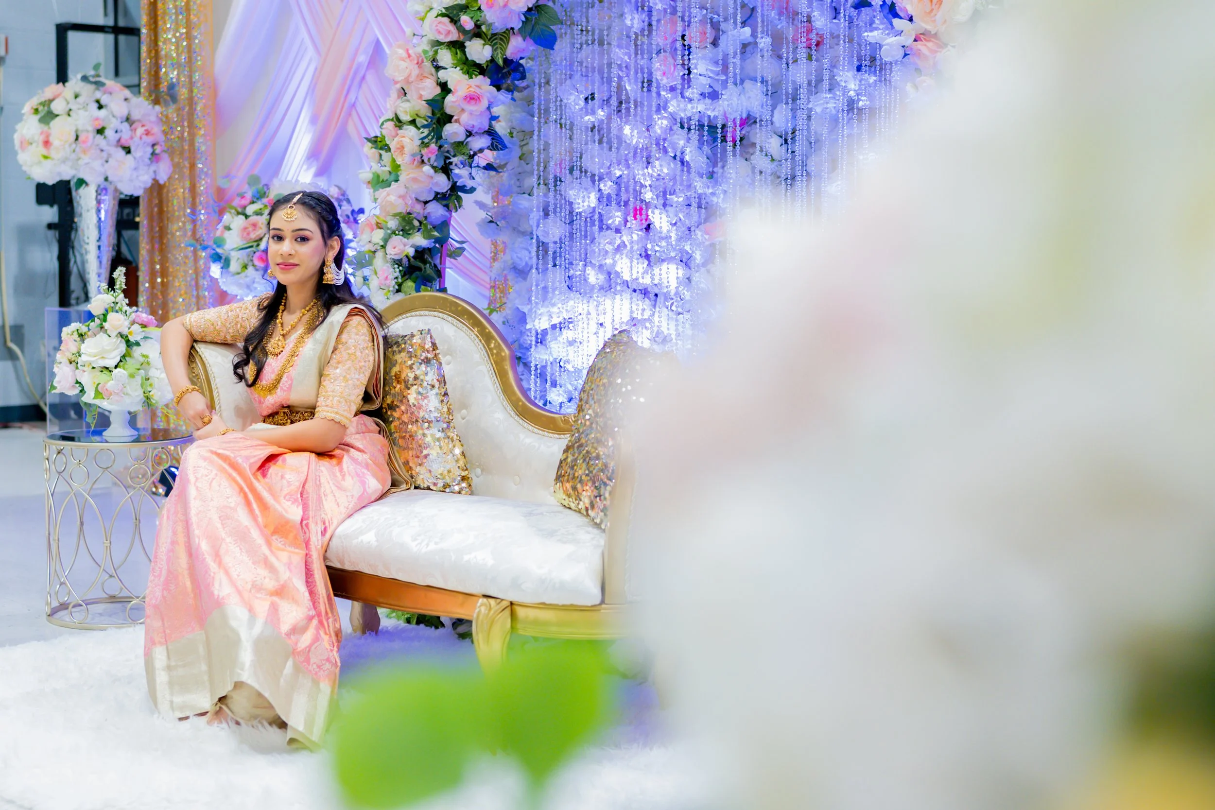 A young woman dressed in traditional pink and gold attire sitting on a decorated sofa at a wedding or celebration. The background features floral arrangements and sparkling decorations, with purple and white lighting.