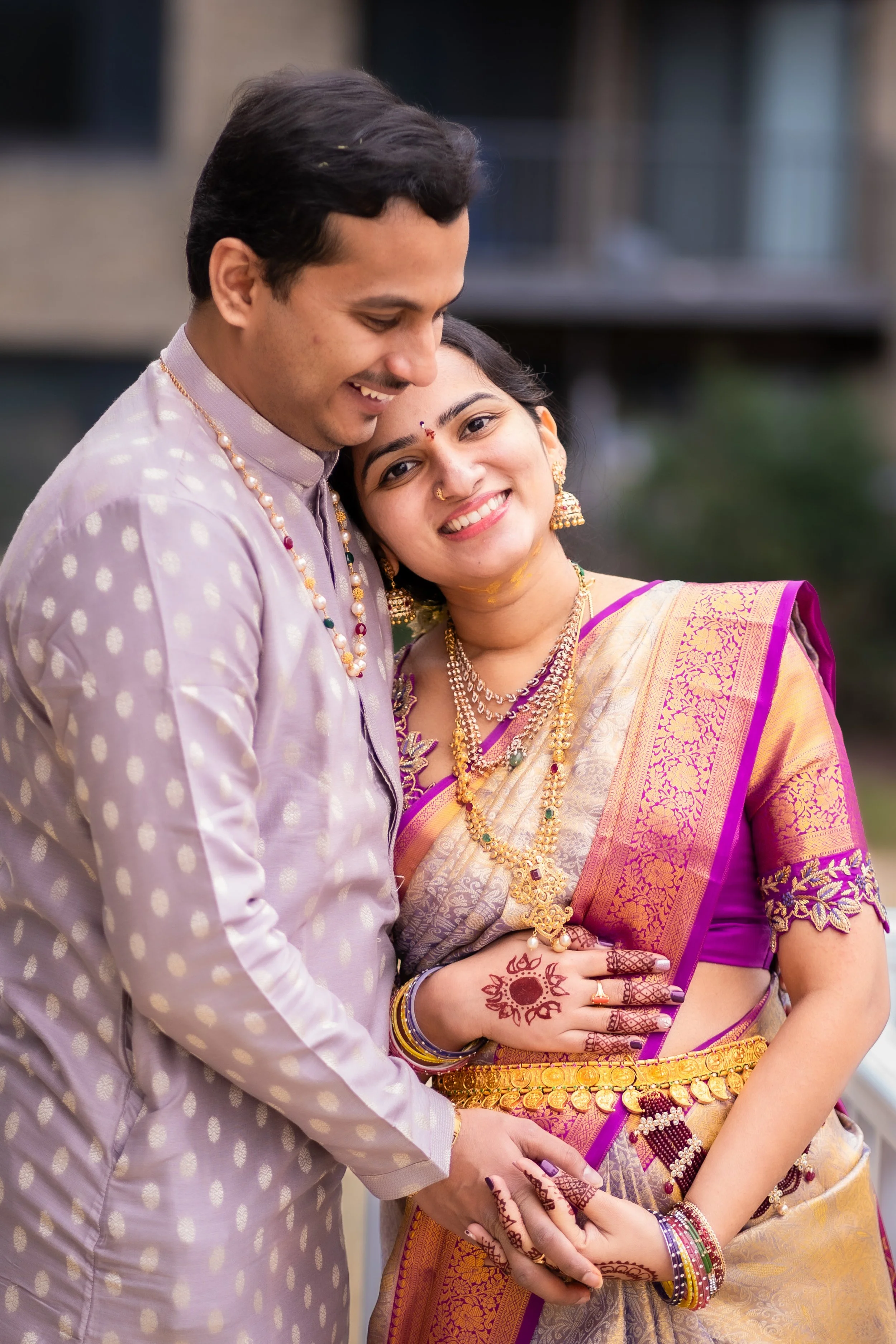 A smiling couple dressed in traditional Indian wedding attire, holding hands with intricate henna on their hands, wearing jewelry and gold coins as accessories.