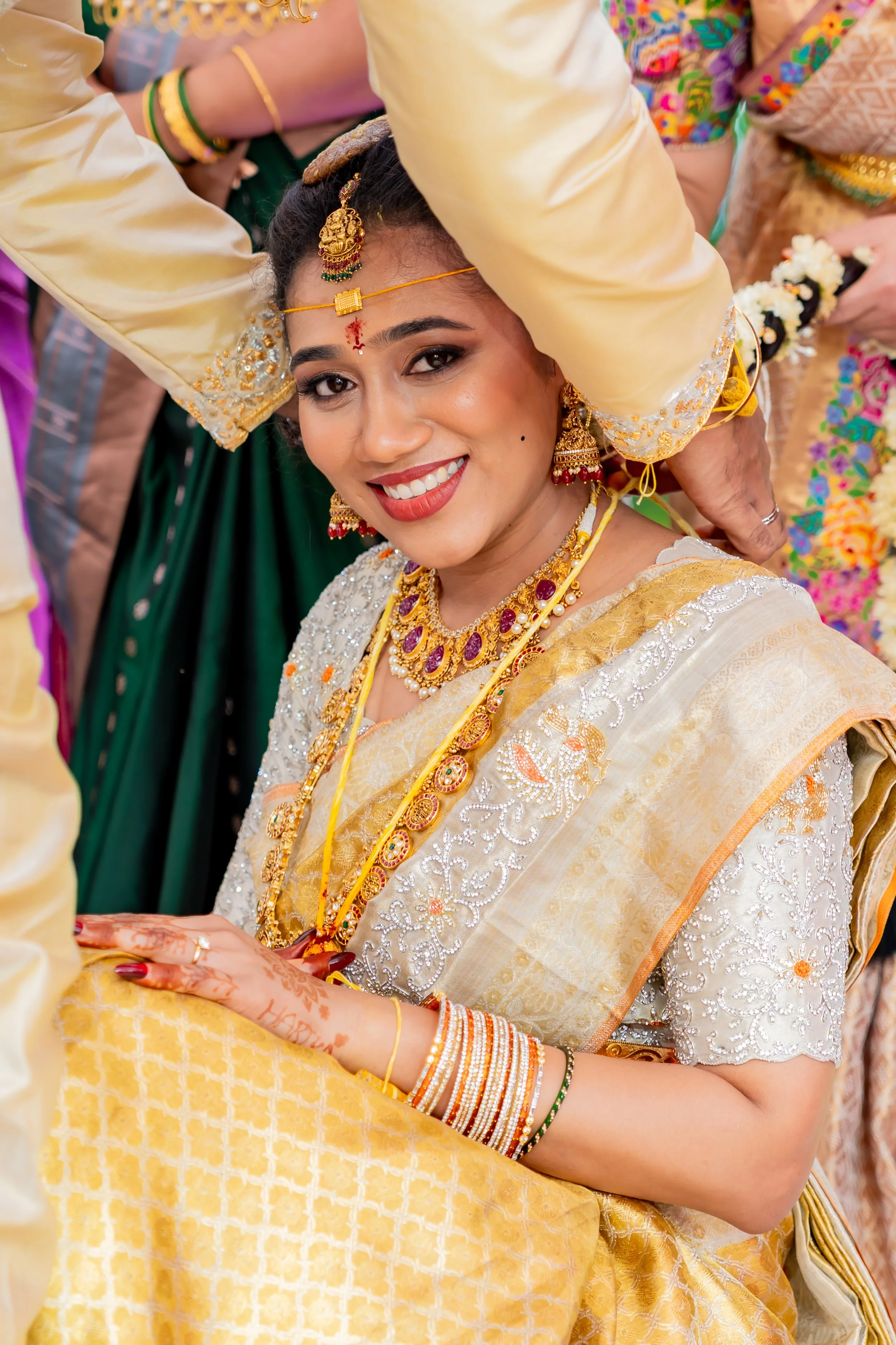 A smiling woman dressed in traditional South Asian bridal attire with jewelry and henna on her hands, participating in a wedding ceremony.