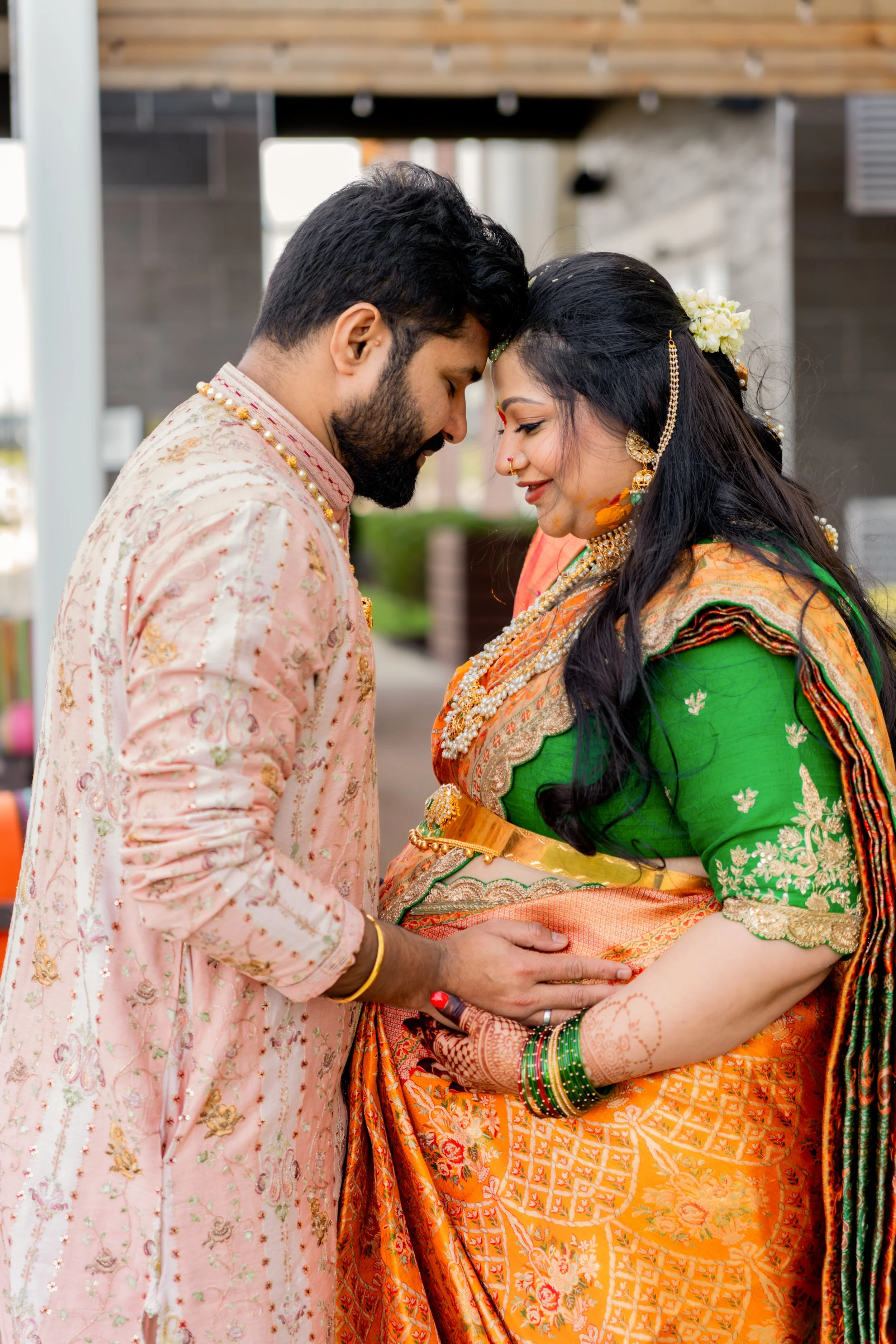 Indian couple dressed in traditional wedding attire sharing a tender moment, with the groom gently holding the bride's belly and both touching foreheads.