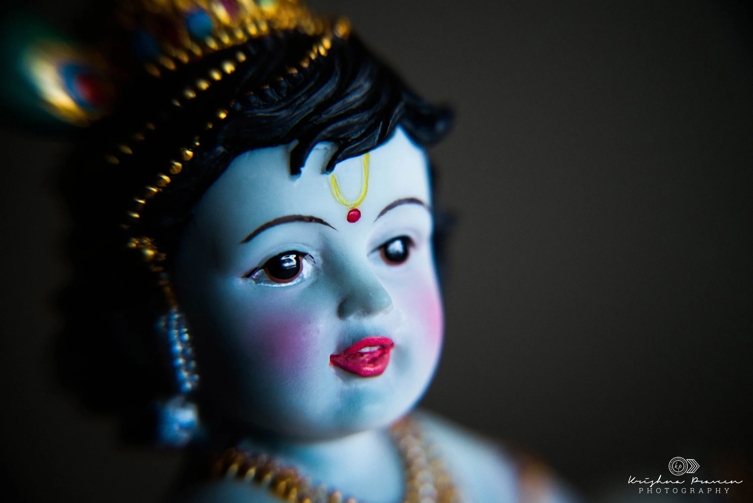 Close-up of a decorative face, likely a traditional sculpture or figurine, with detailed black hair, a red bindi on the forehead, expressive eyes, and pink lips, adorned with colorful jewelry.