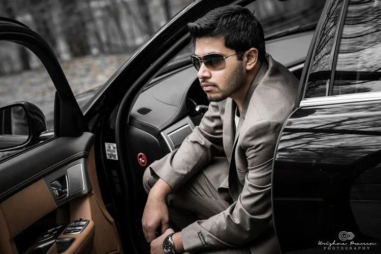 A man in a gray suit and sunglasses sits on the edge of a black car with the door open, looking to the side.