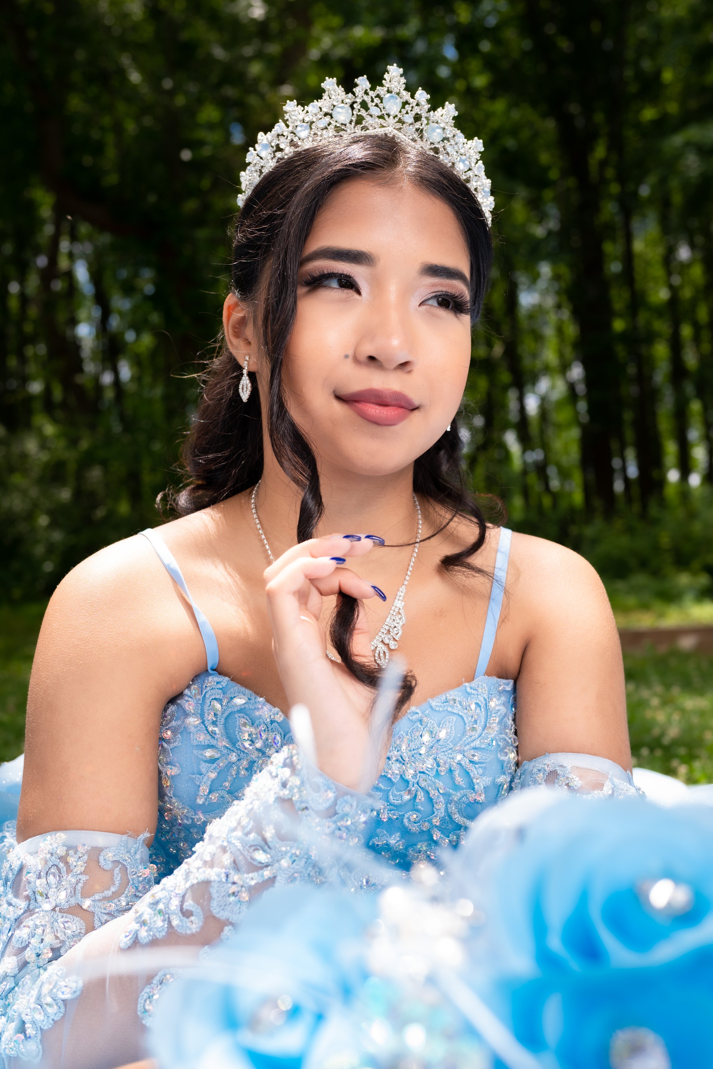 Close-up of a young woman with dark hair, wearing a silver tiara and a light blue dress with lace and embroidery, outdoors with blurred green trees in the background.