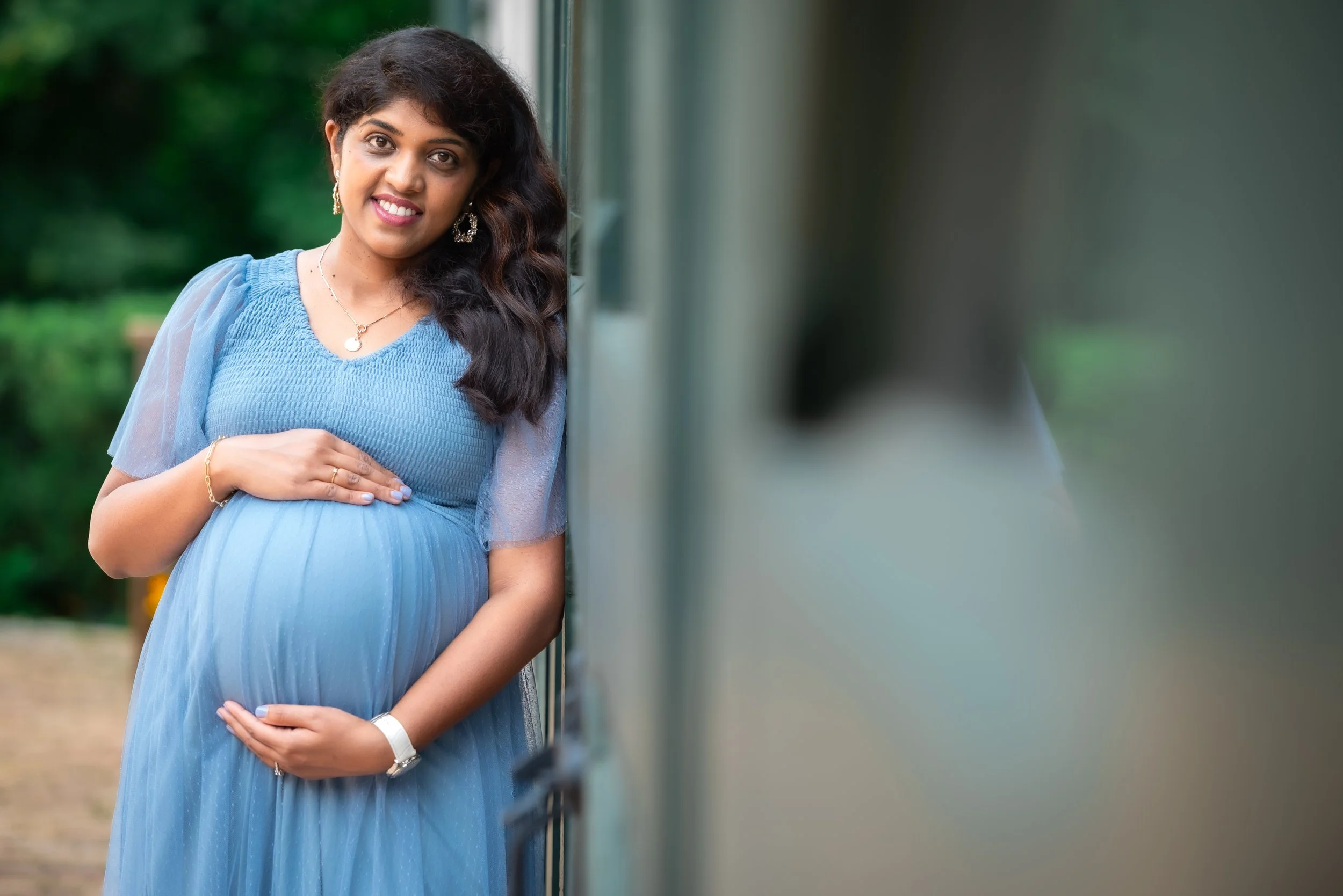 A pregnant woman smiling, wearing a blue dress and jewelry, standing outdoors near a reflective surface in a natural, green environment.