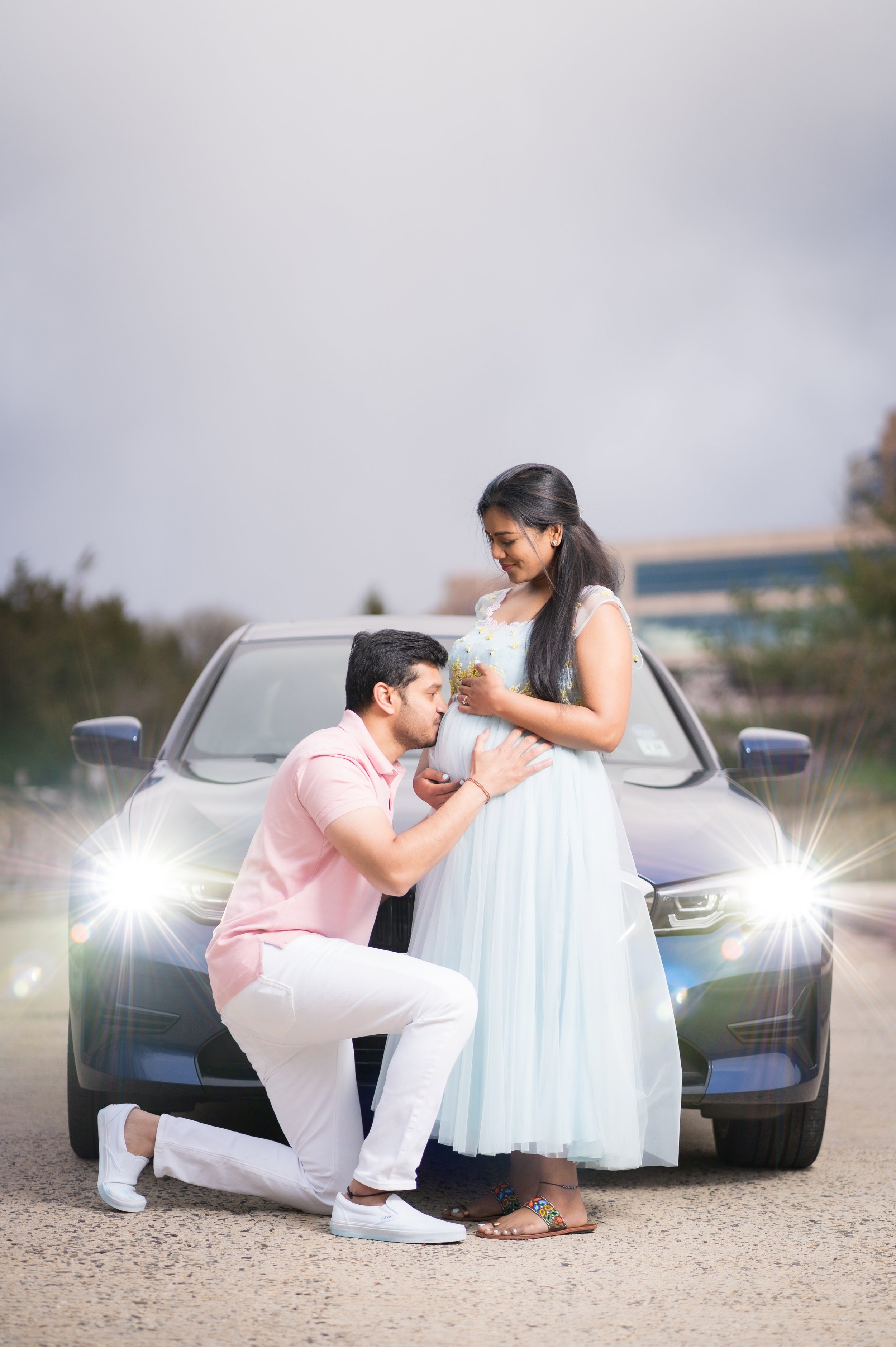 A man kneeling on one knee and holding a pregnant woman by the waist in front of a car, with their heads close, outdoors on a cloudy day.