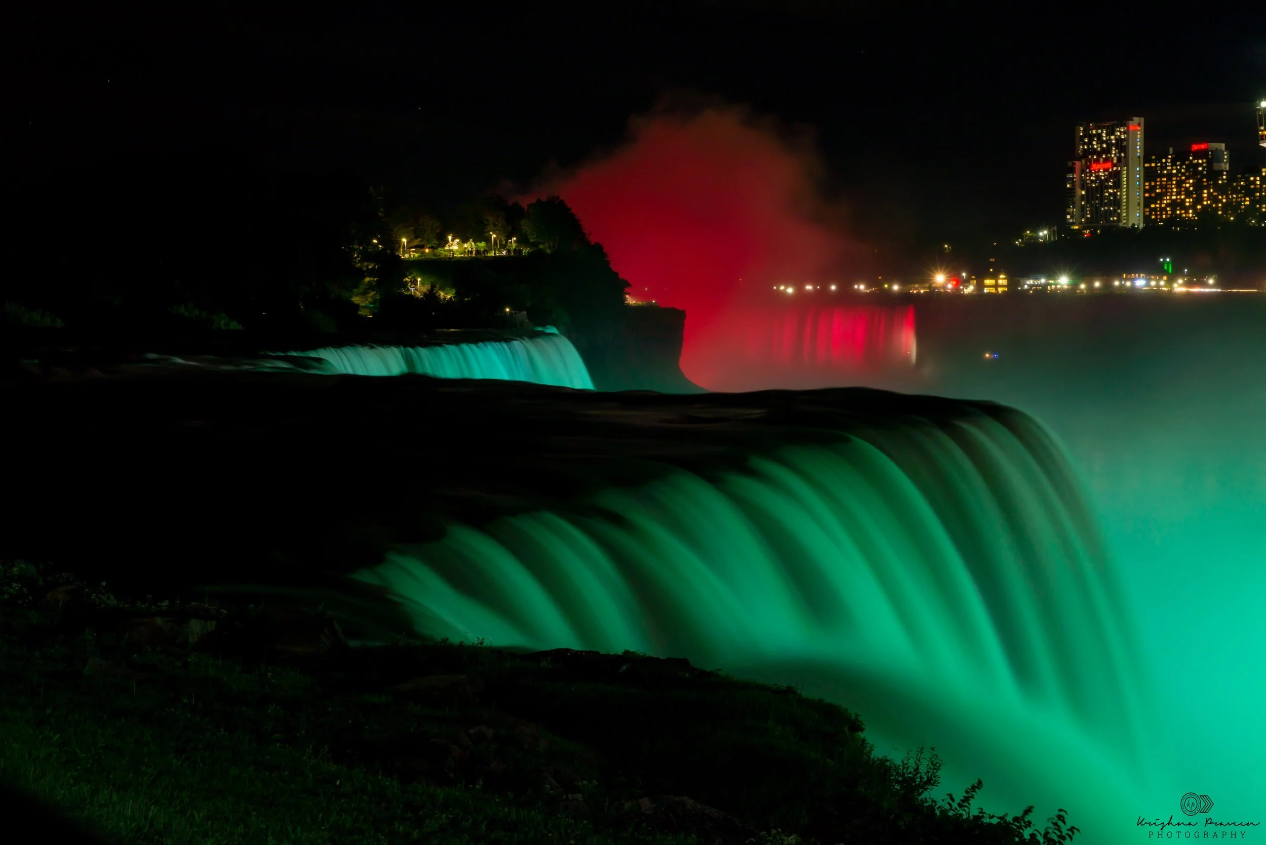 Night view of Niagara Falls illuminated with green and red lights, with water spray rising into the night sky and a city skyline with lit buildings in the background.