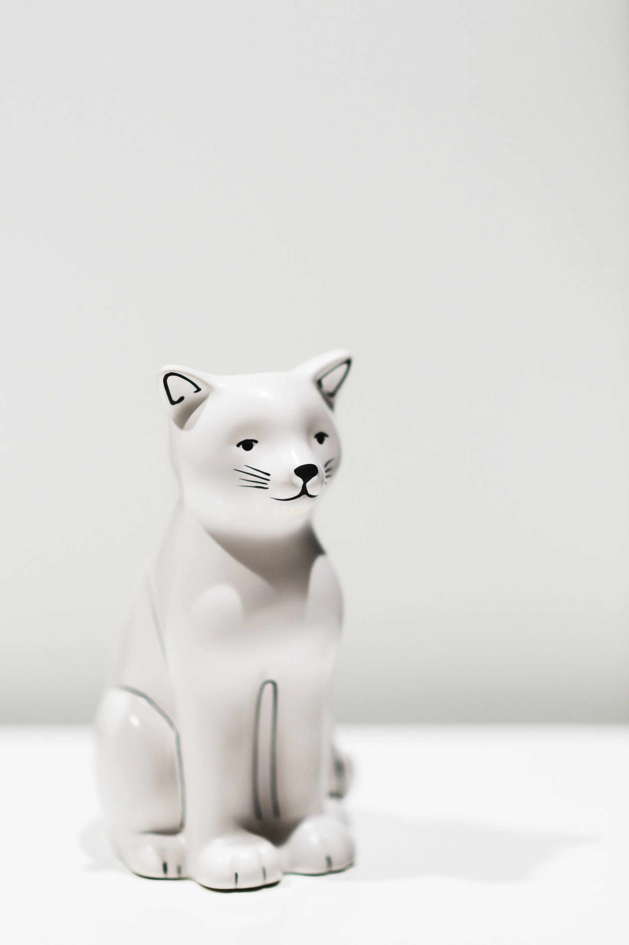 White ceramic cat figurine with black line drawings of a face, standing on a plain white surface against a white background.