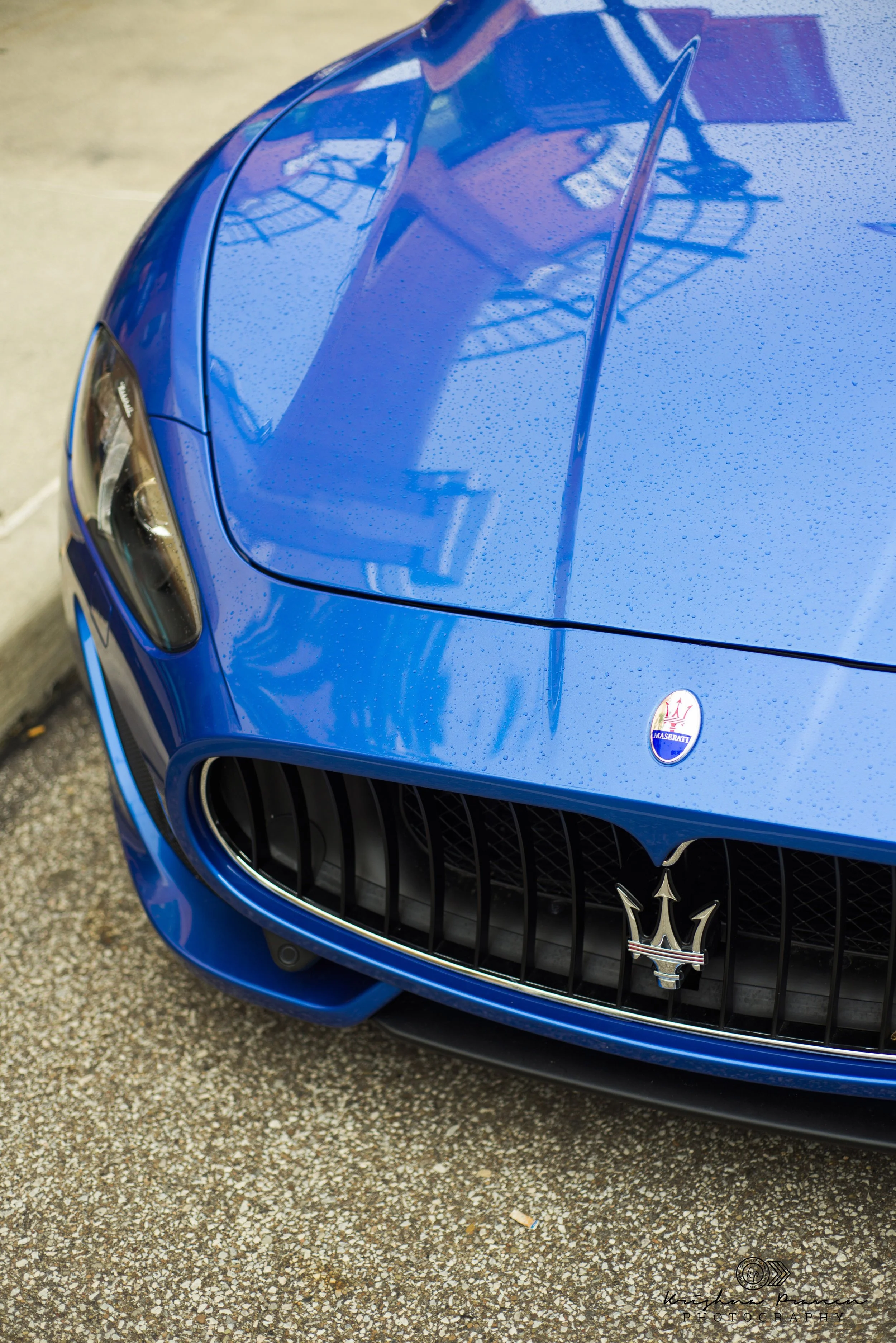 Close-up of front part of a blue Maserati sports car with water droplets on the hood, showing the Maserati logo and a black grill with vertical slats.
