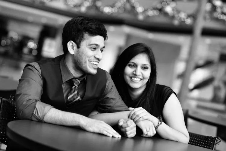 A man and woman sitting at a table, smiling and leaning on each other in an indoor setting.