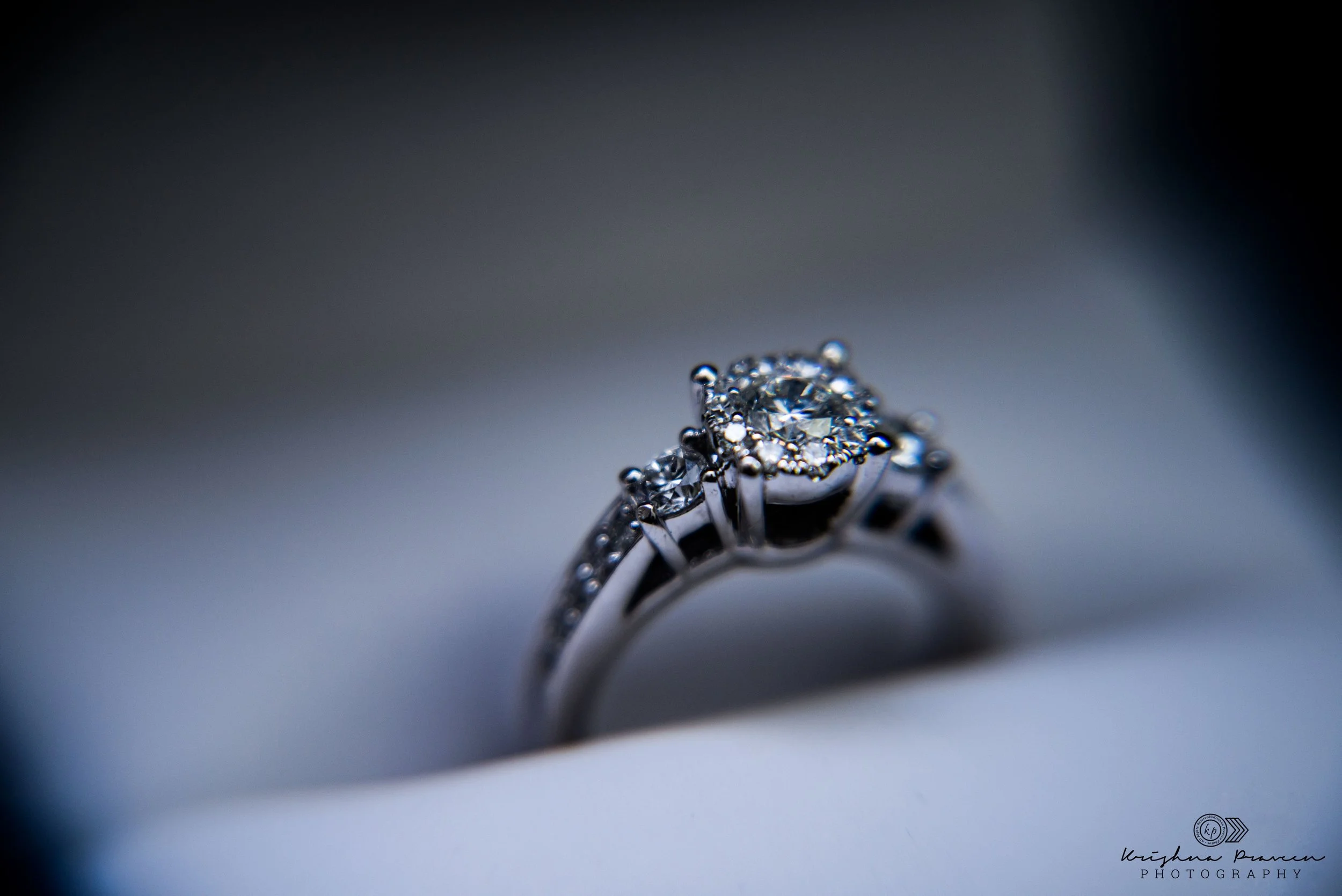 Close-up of a diamond engagement ring with multiple smaller diamonds on a silver band, against a blurred background.