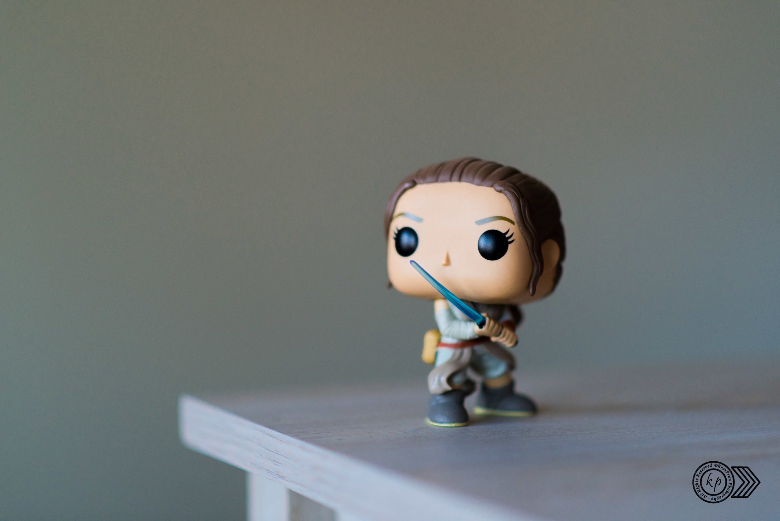 A stylized figurine of a female character with brown hair, holding a blue sword, dressed in beige and brown clothing, standing on a white platform against a plain background.