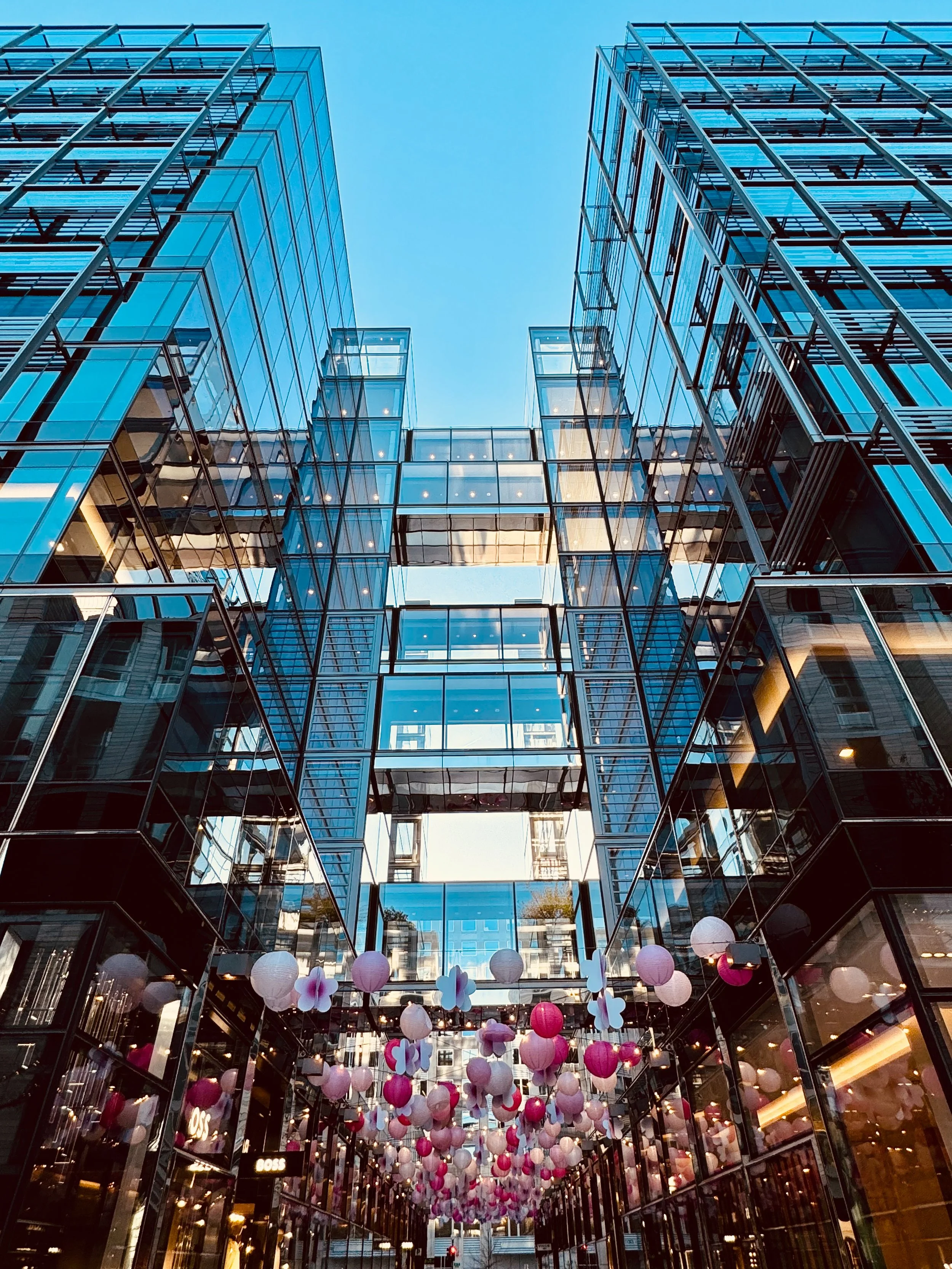 Looking up at the glass-fronted buildings in a city with hanging pink and white paper lanterns and decorations.