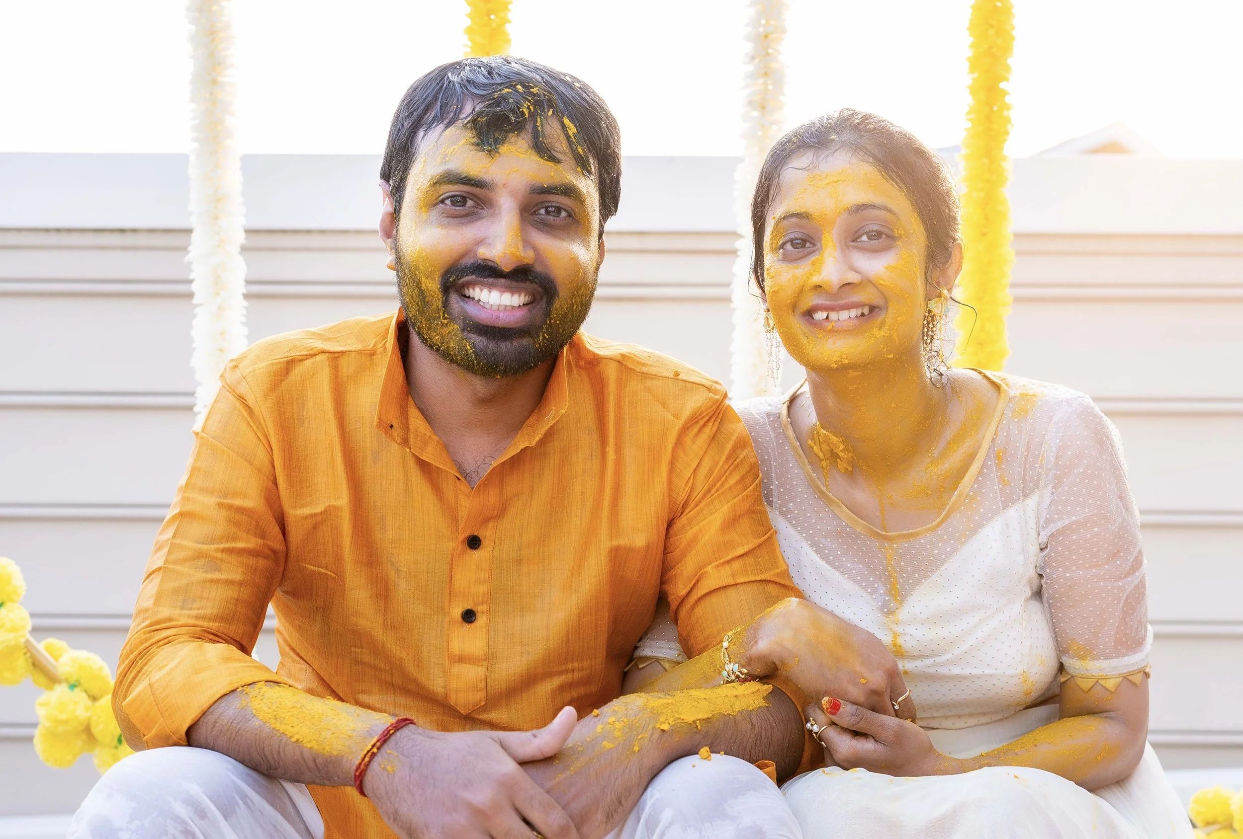 A happy couple celebrating a traditional Indian festival, covered in yellow turmeric powder, sitting outdoors with floral decorations behind them.