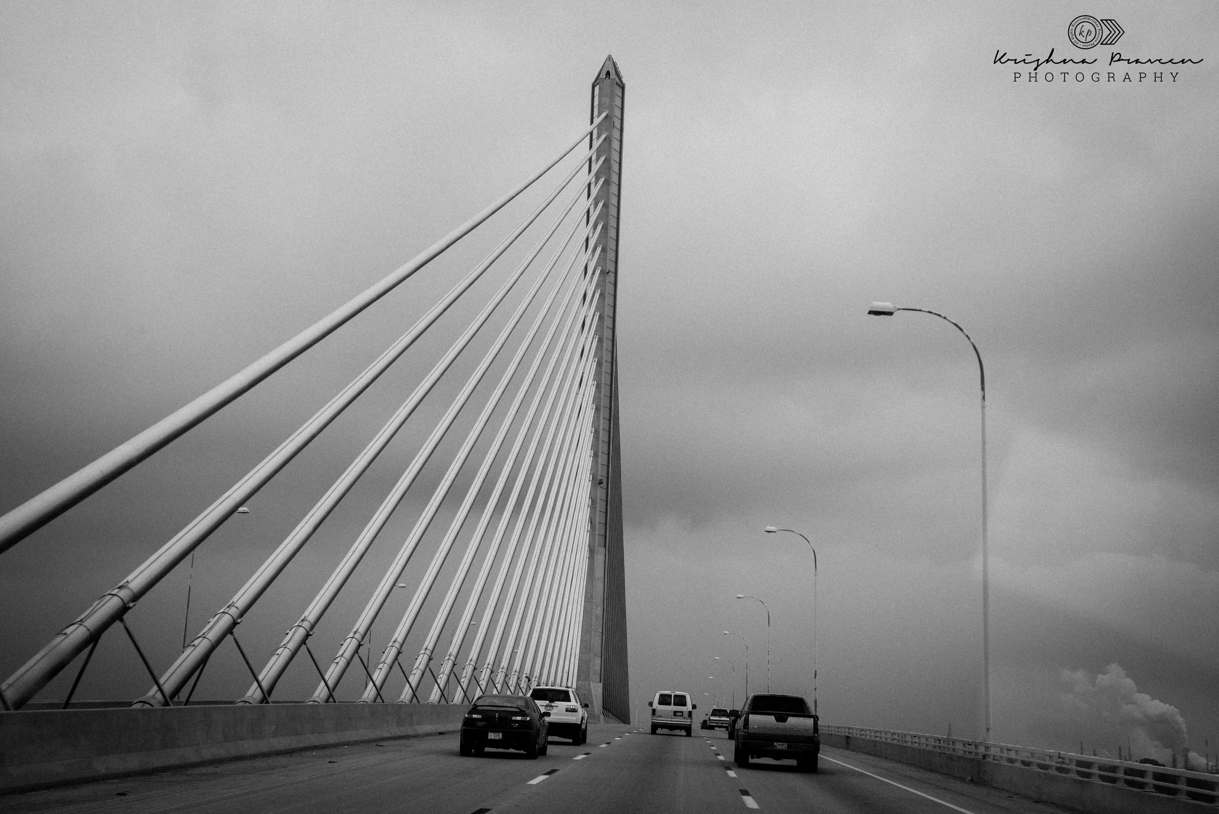 Black and white photo of cars driving on a bridge with a tall cable-stayed tower and streetlights along the edge of the roadway.