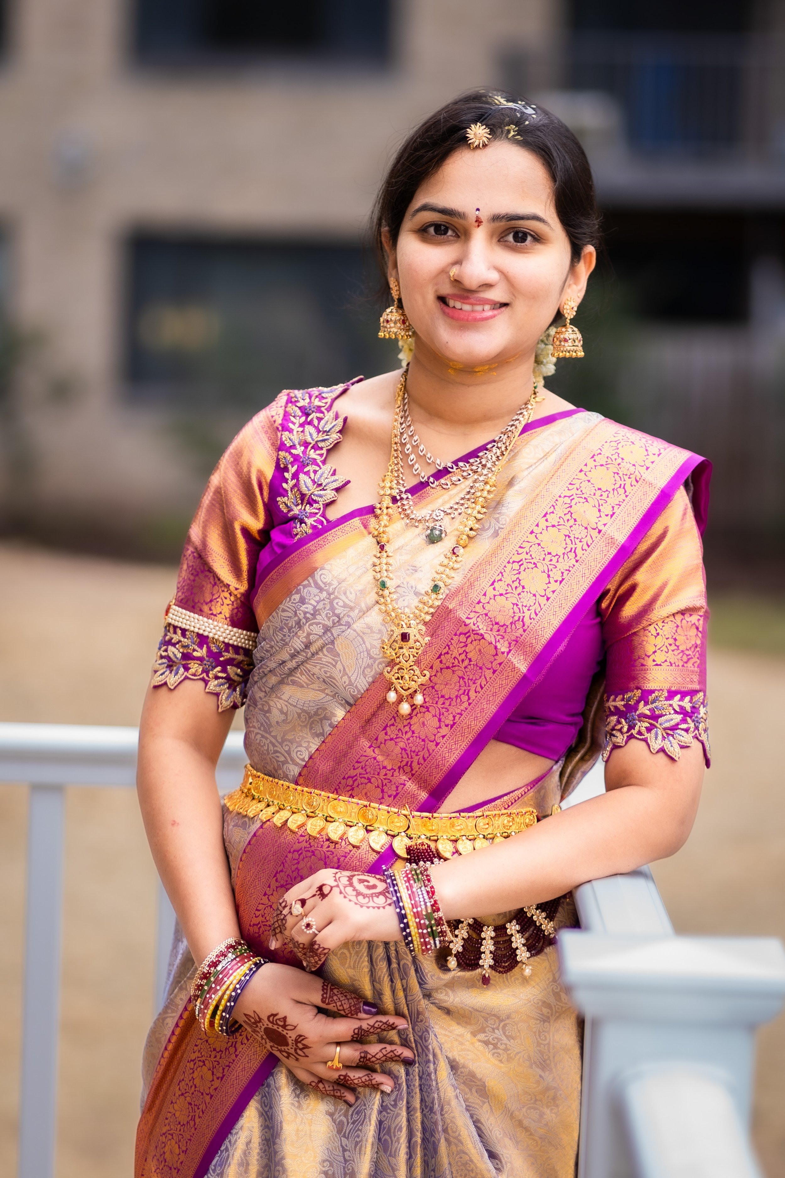 A woman in traditional Indian attire smiling outdoors, wearing a purple and gold saree with jewelry including necklaces, earrings, bangles, and a nose ring.