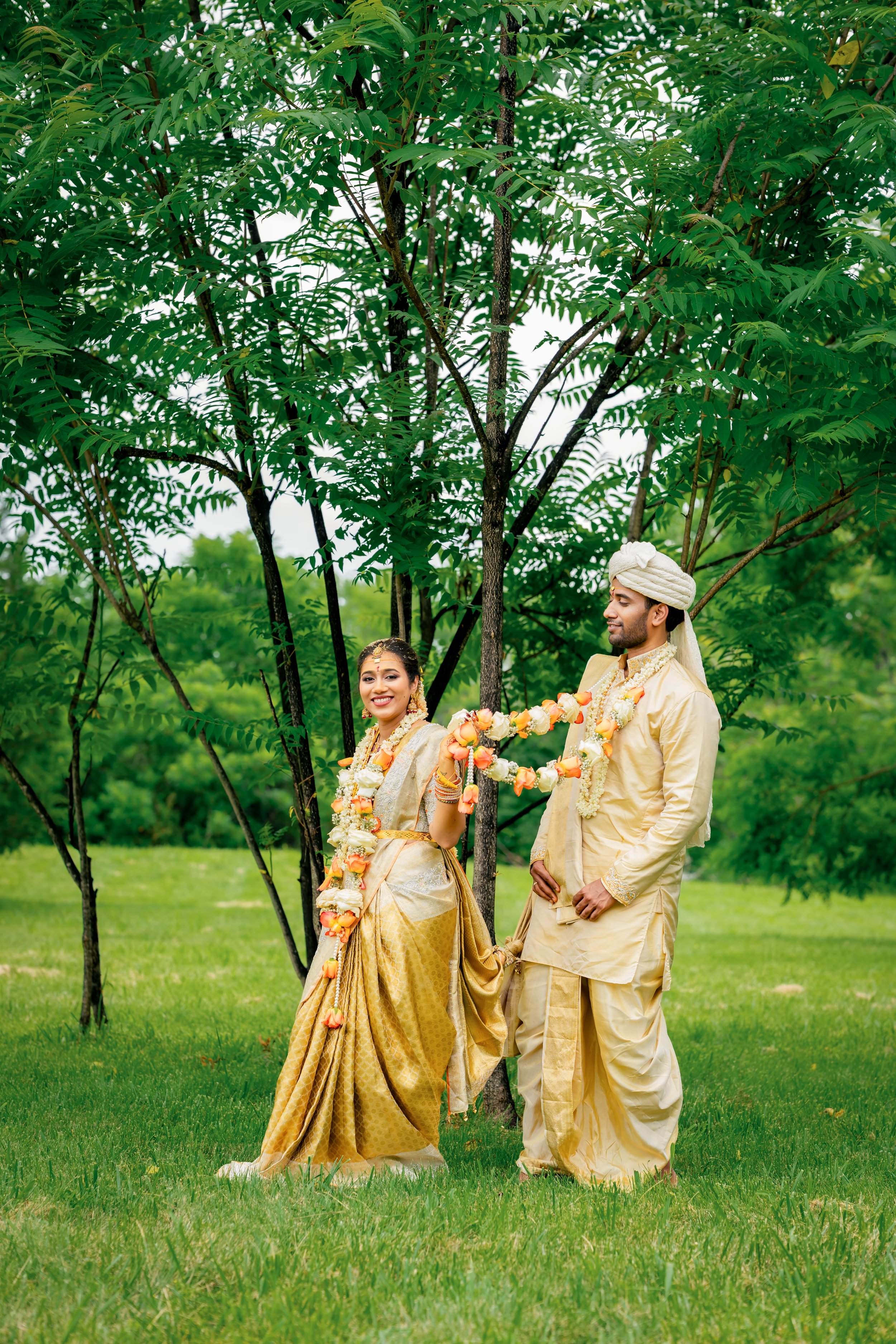 A newlywed Indian couple dressed in traditional wedding attire standing outdoors under a large green tree, with lush greenery in the background.