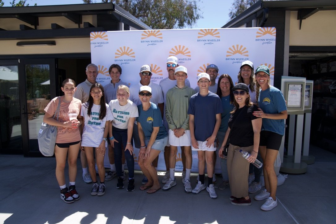 Group photo of people in front of a Brynn Wheeler Foundation banner outside a building on a sunny day.