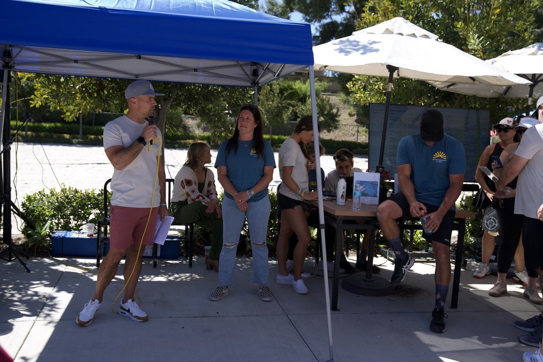 A group of people gathered outdoors under tents, with one man speaking into a microphone, others sitting or standing nearby, some with water bottles, on a sunny day with trees in the background.