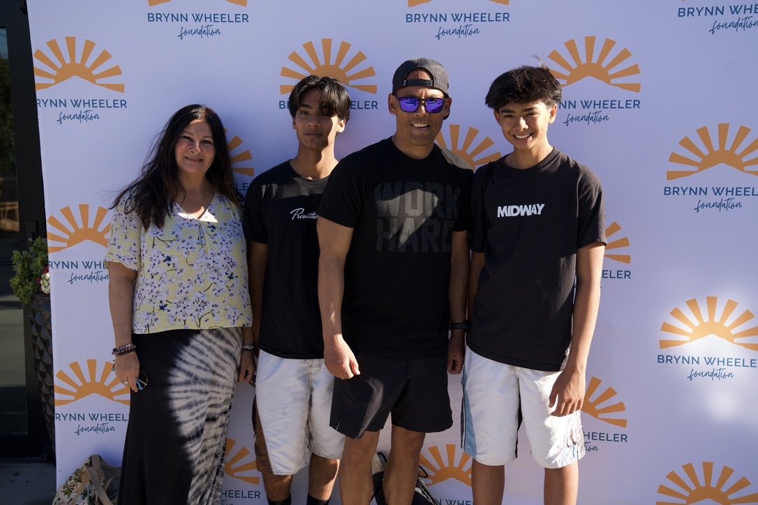 Four people standing in front of a Brynn Wheeler Foundation backdrop, including a woman and three young men, all smiling. The woman has long dark hair, wearing a floral blouse and patterned skirt. The second person is a young man with dark hair and a