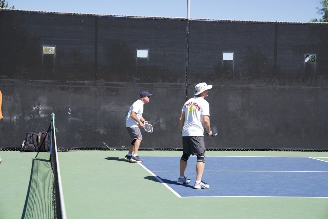 Two men playing pickleball on an outdoor court, with a black fence in the background and a backpack resting on the ground.