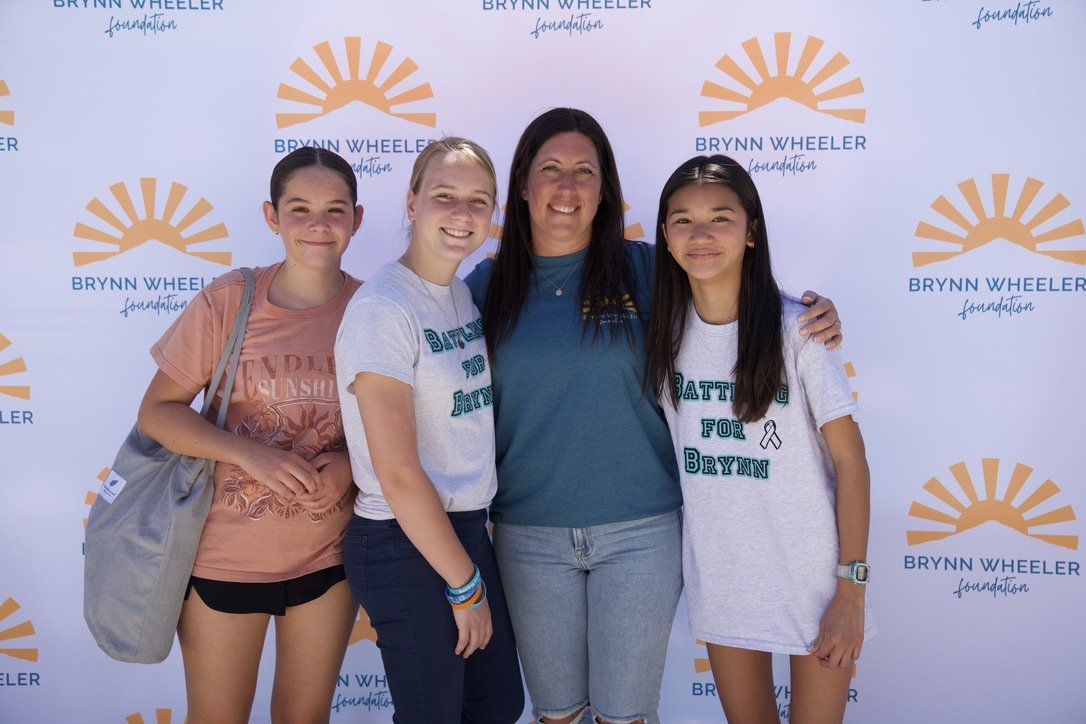 Four girls and one woman standing in front of a backdrop with the Bryn Wheeler Foundation logo, smiling at the camera. The woman is in the center, wearing a teal shirt. The girls are wearing T-shirts with messages supporting Bryn and a pink ribbon, i