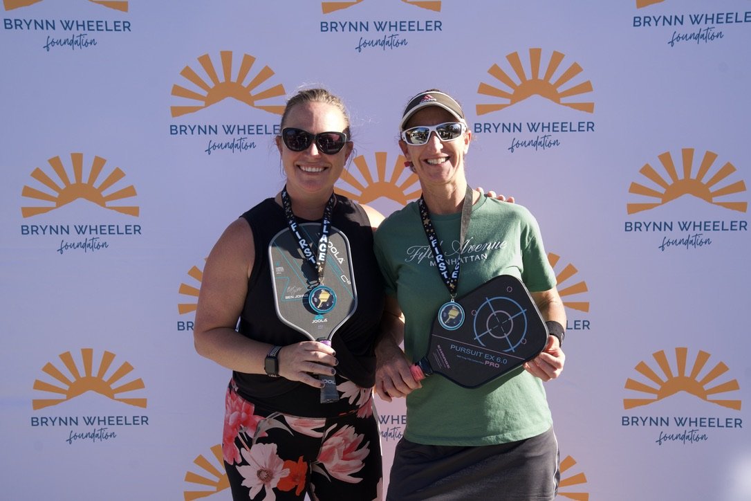 Two women in athletic attire smiling and posing together, each holding a pickleball paddle, standing in front of a backdrop with 'Brynn Wheeler Foundation' logo and sunburst design.