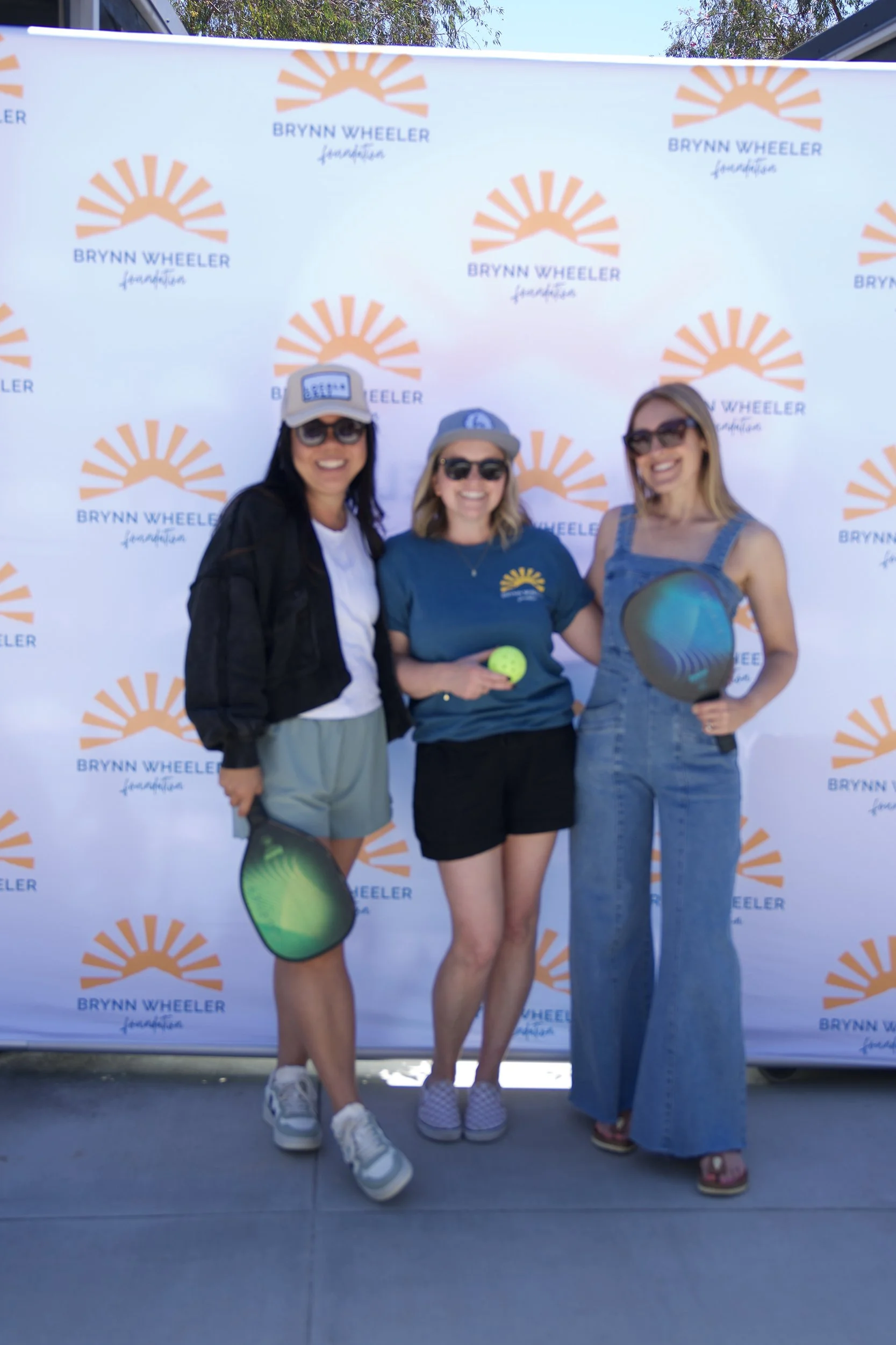 Three women standing together in front of a backdrop with the Brynn Wheeler Foundation logo. They are smiling and holding pickleball paddles and a tennis ball. One woman is wearing sunglasses, a hat, a jacket, and shorts; the middle woman is wearing 