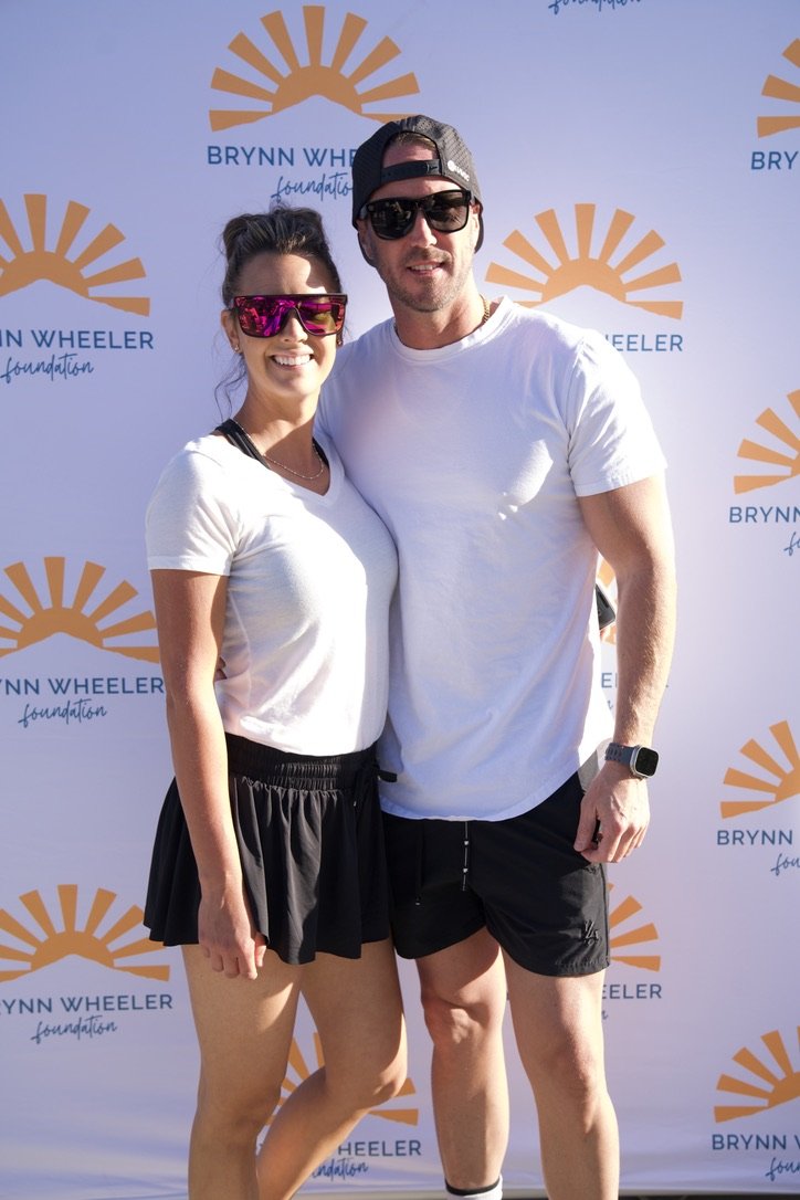 A smiling woman and man standing together outdoors, wearing casual athletic clothing and sunglasses, in front of a backdrop with the Brynn Wheeler Foundation logo featuring an orange sunburst.