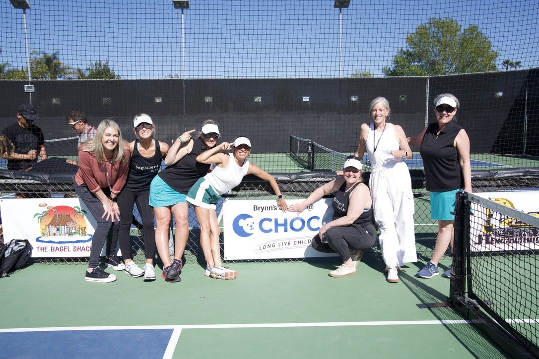 Group of seven women and girls posing on a tennis court, smiling, with a chain-link fence and a black windscreen behind them. Two of them wear white visors, and all are dressed in sporty or casual attire. The sun is shining, and there are trees and a