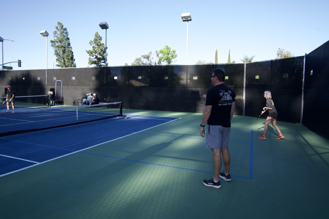People playing pickleball on an outdoor court with a black wall and surrounding trees.