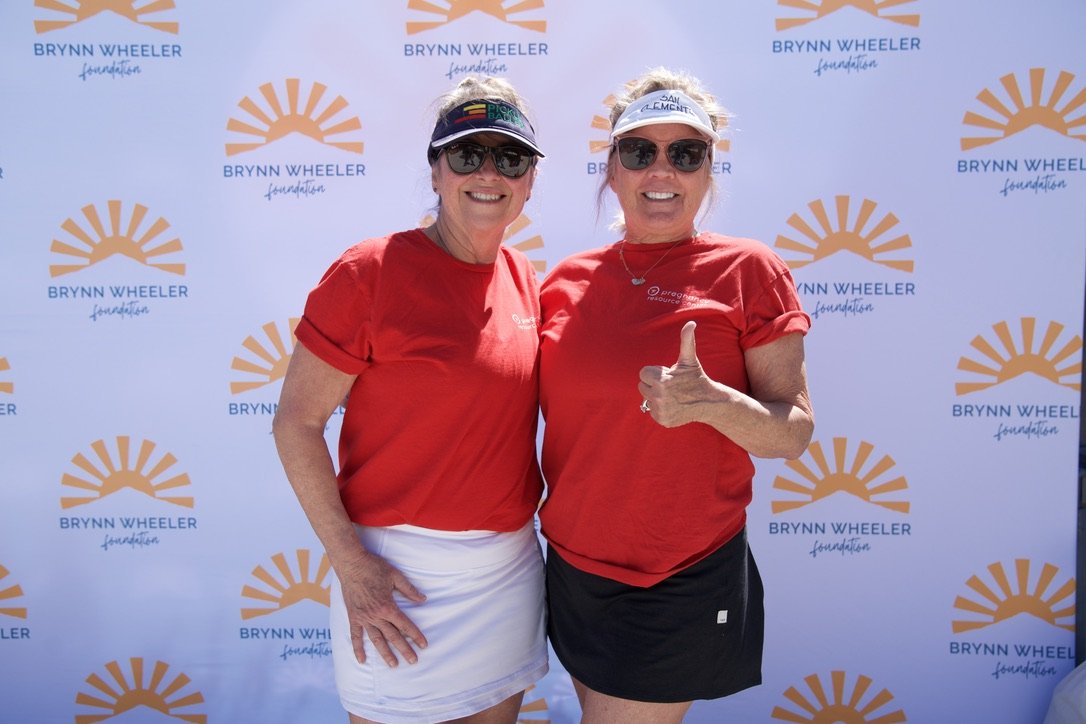 Two women in red shirts and sunglasses smiling and posing in front of a Brynn Wheeler Foundation backdrop. One woman is giving a thumbs-up.