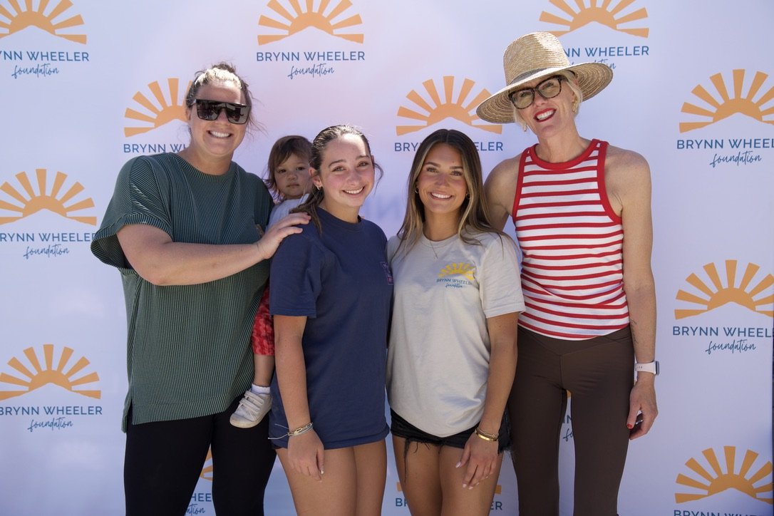 Group of five women and a young girl posing for a photo in front of a Brynn Wheeler Foundation backdrop. They are smiling and wearing casual summer clothing.