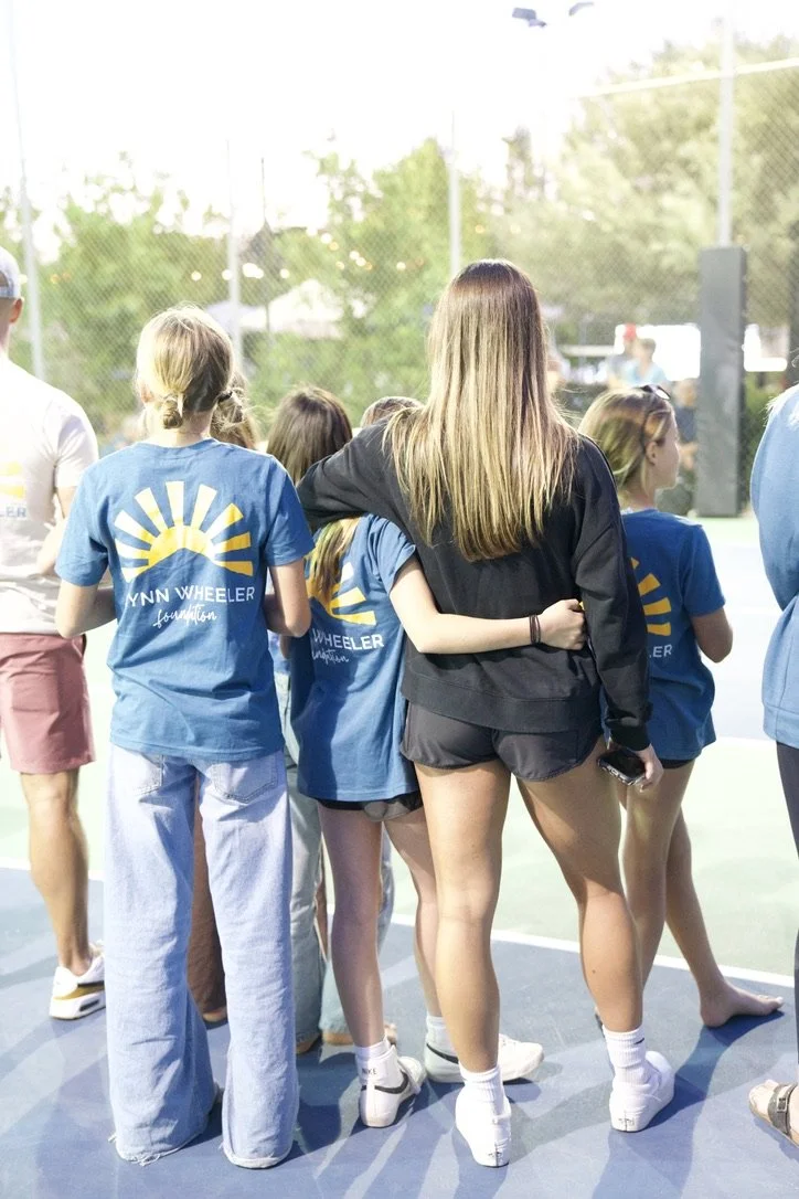 Group of young girls and women standing together on an outdoor tennis court, some wearing blue T-shirts and shorts, near the net, with trees and a fence in the background during daytime.