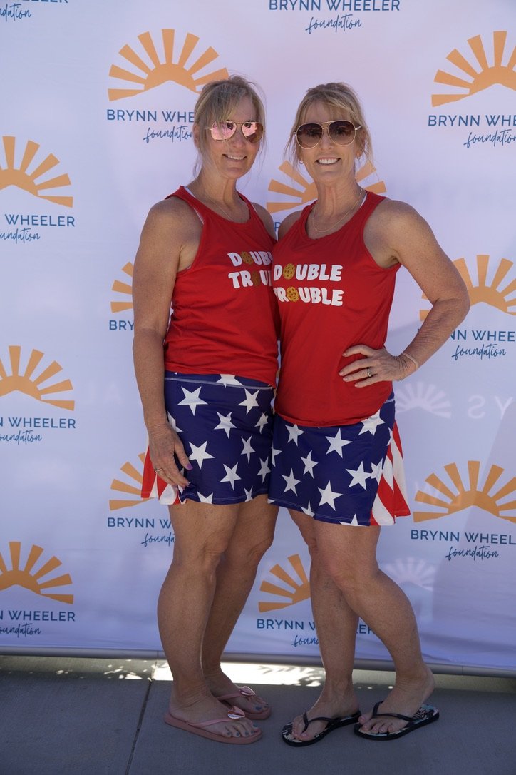 Two women dressed in patriotic attire with red tops and star-spangled shorts stand in front of a BLYNN WHEELER foundation backdrop, smiling at the camera.
