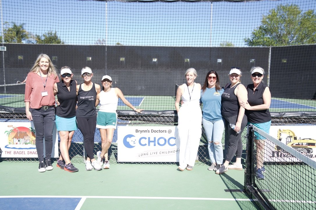Group of women standing on a tennis court, smiling, with a tennis net and banners in the background.