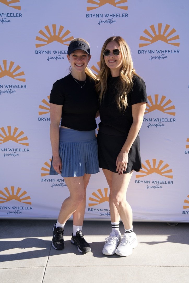 Two young women standing together in front of a Brynn Wheeler Foundation backdrop, smiling and posing for the photo, both wearing black t-shirts and athletic shoes.